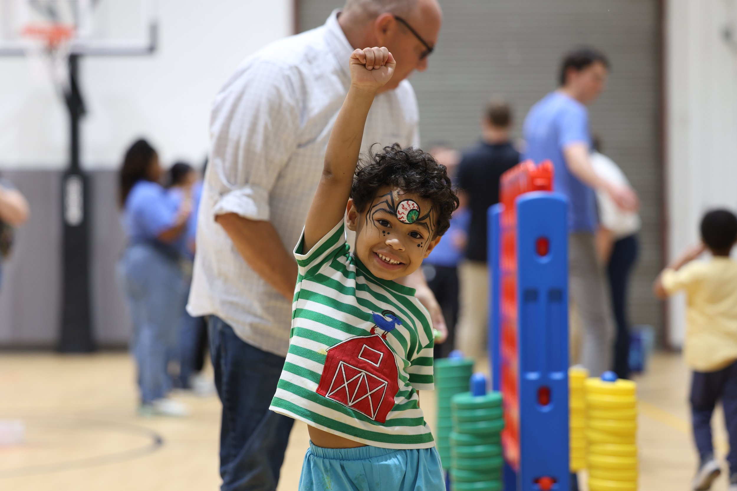A young boy with face paint and a big smile, raising his arm in a gesture of victory or excitement, at an indoor event or playground. A man stands behind him, and other children and adults are visible in the background.