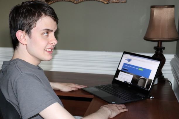 A young person with short brown hair sitting at a wooden desk, using a laptop, with a beige lamp and a decorative mirror on the wall behind.