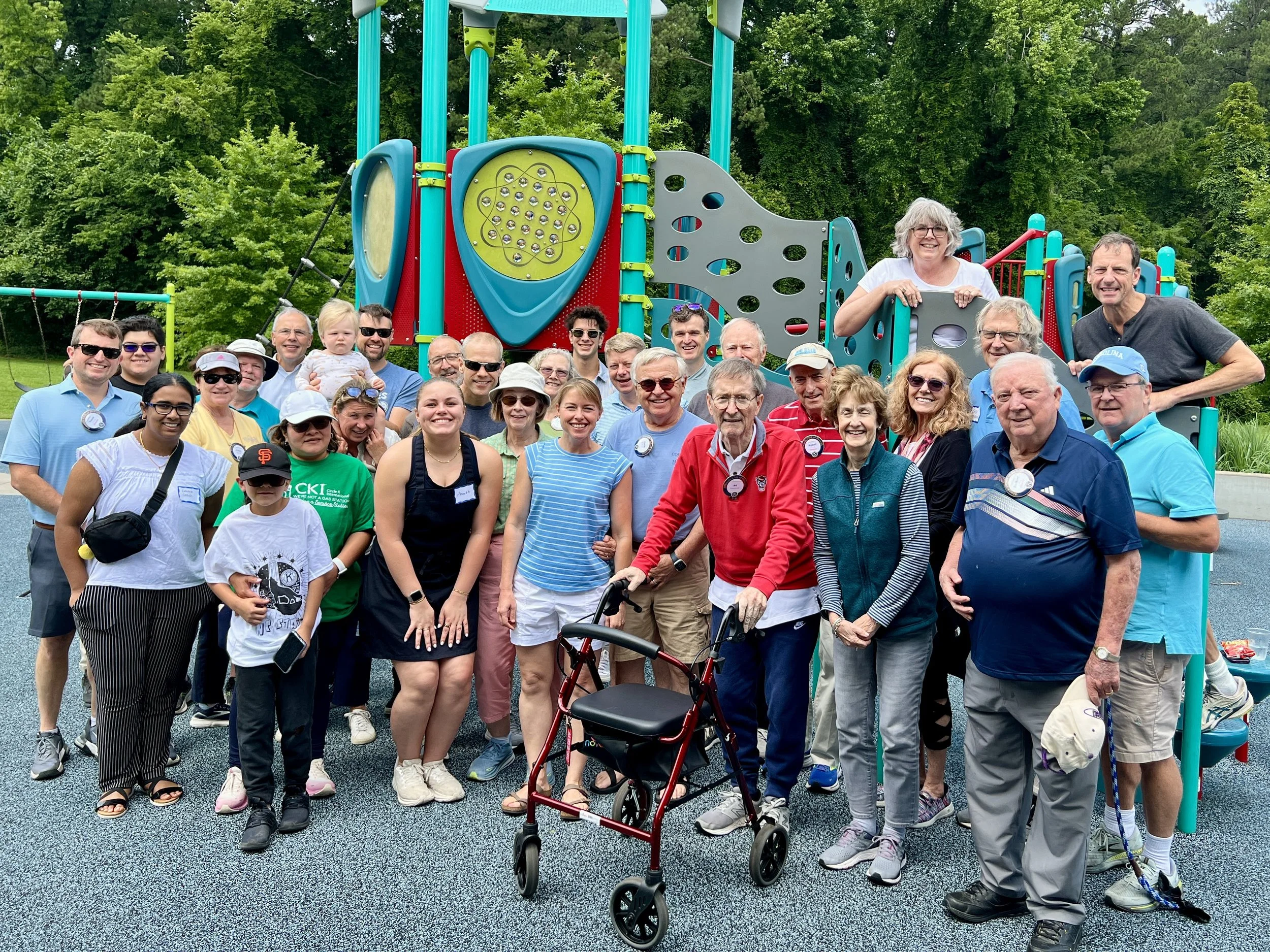 Group of people, including children and elderly, gathered outdoors in front of a colorful playground, smiling for a photo.