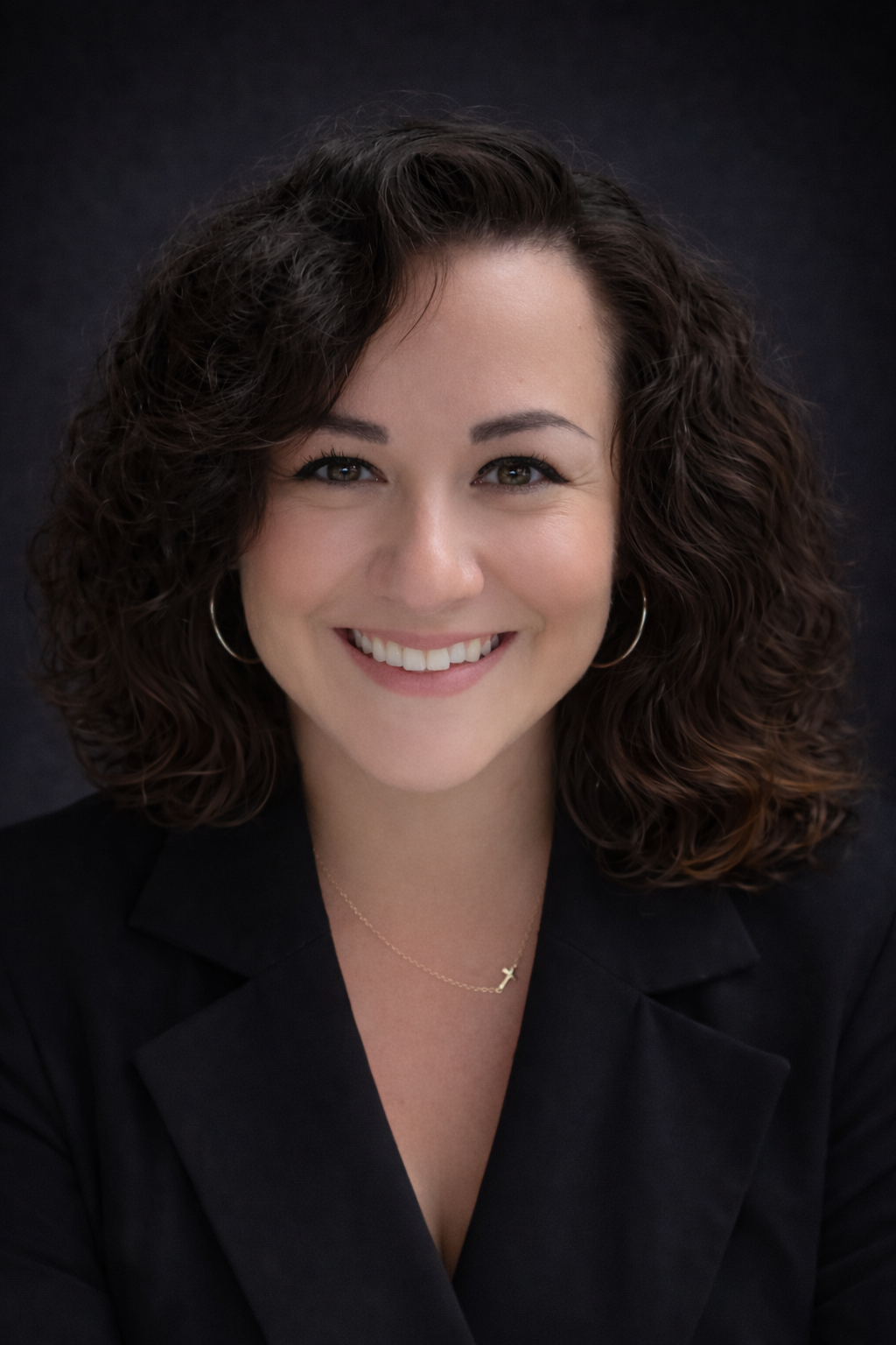 A young woman with curly brown hair smiling at the camera. She is wearing hoop earrings, a delicate cross necklace, and a black blazer against a dark background.