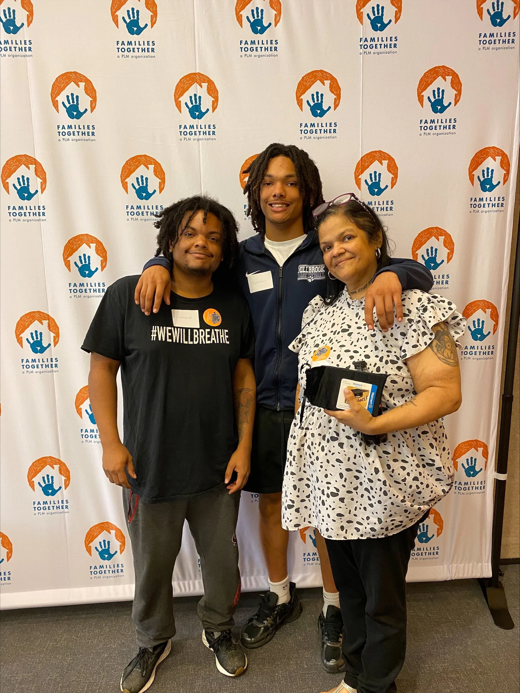 Three people standing together in front of a backdrop with the 'Families Together' logo. The person on the left wears a black T-shirt that says '#WEWILLBREATHE'. The person on the right wears a white dress with black spots and holds a container. The 