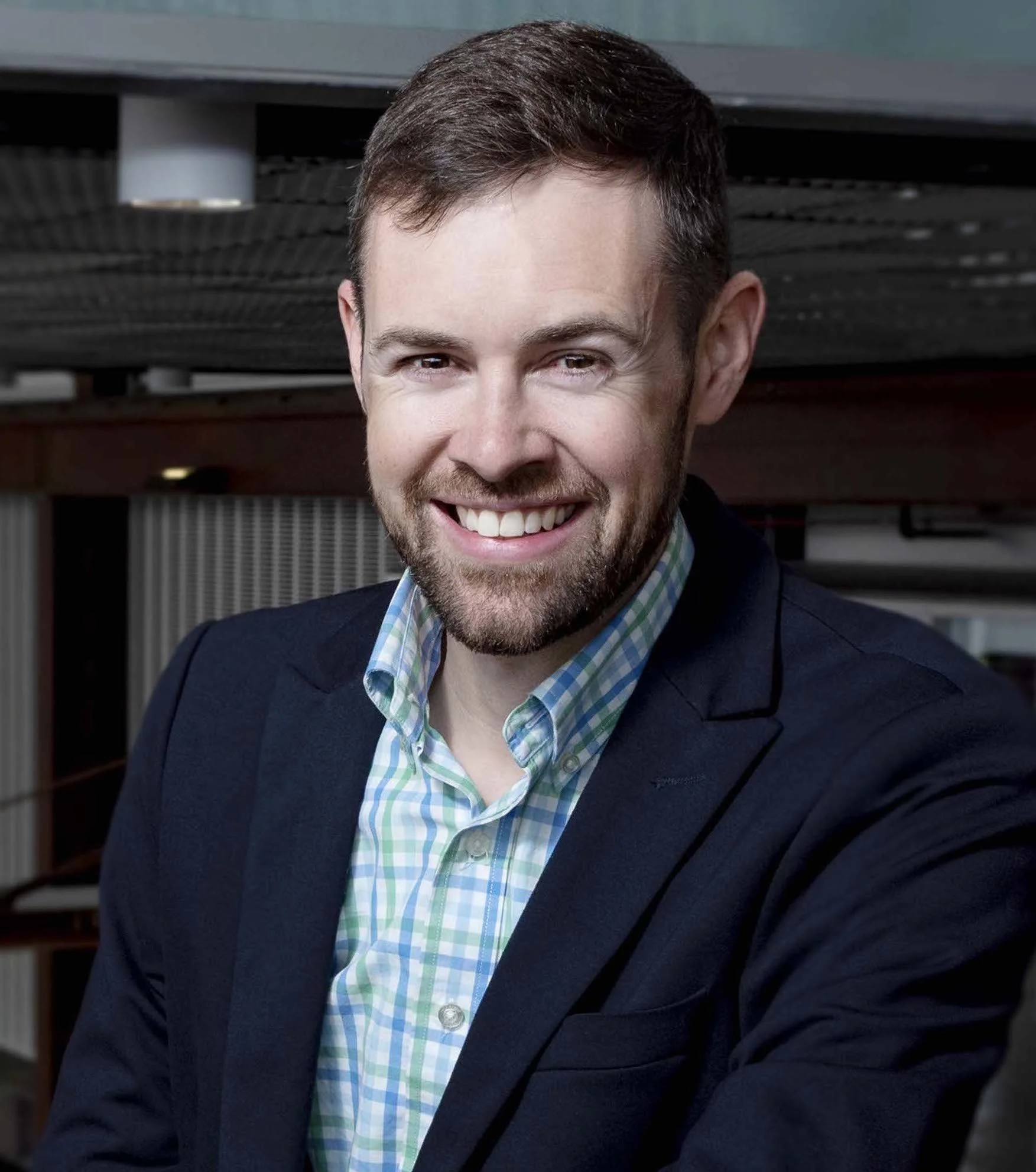 A smiling man in a blue blazer and checkered shirt in an indoor setting.