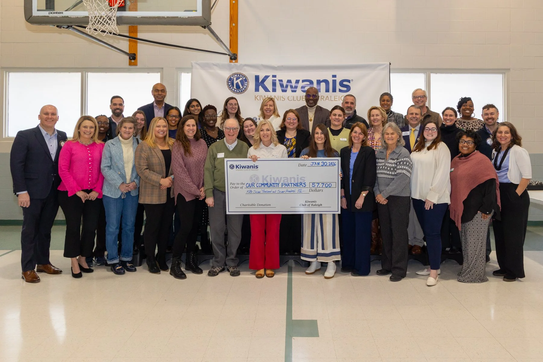 Group of people posing with a large check for $57,700 made out to 'Our Community Partners' at a Kiwanis event in a gymnasium, with a basketball hoop and a banner that reads 'Kiwanis Club Raleigh' in the background.