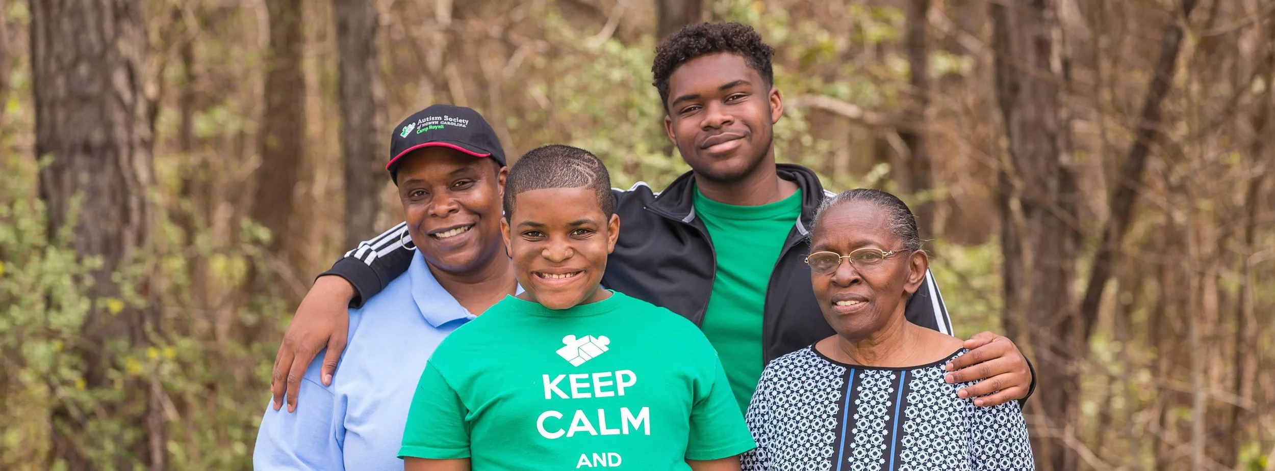 A group of four people, including a young girl and three adults, standing close together outdoors in a wooded area. They are smiling and appear happy, with some members having their arms around each other. The girl wears a green shirt with message, the woman on the left wears a black cap and blue shirt, and the man on the right wears a black jacket over a green shirt, while the elderly woman has glasses and a patterned blouse.