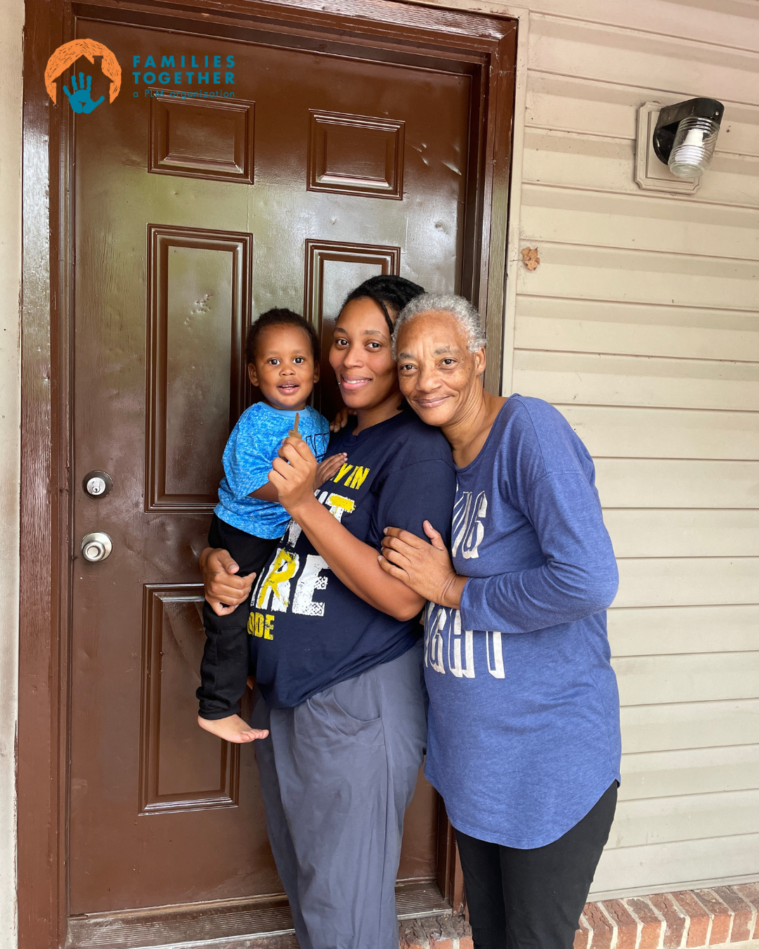 Three people standing in front of a door, hugging and smiling. A young woman is holding a small child, and an elderly woman is hugging the young woman. The door has a 