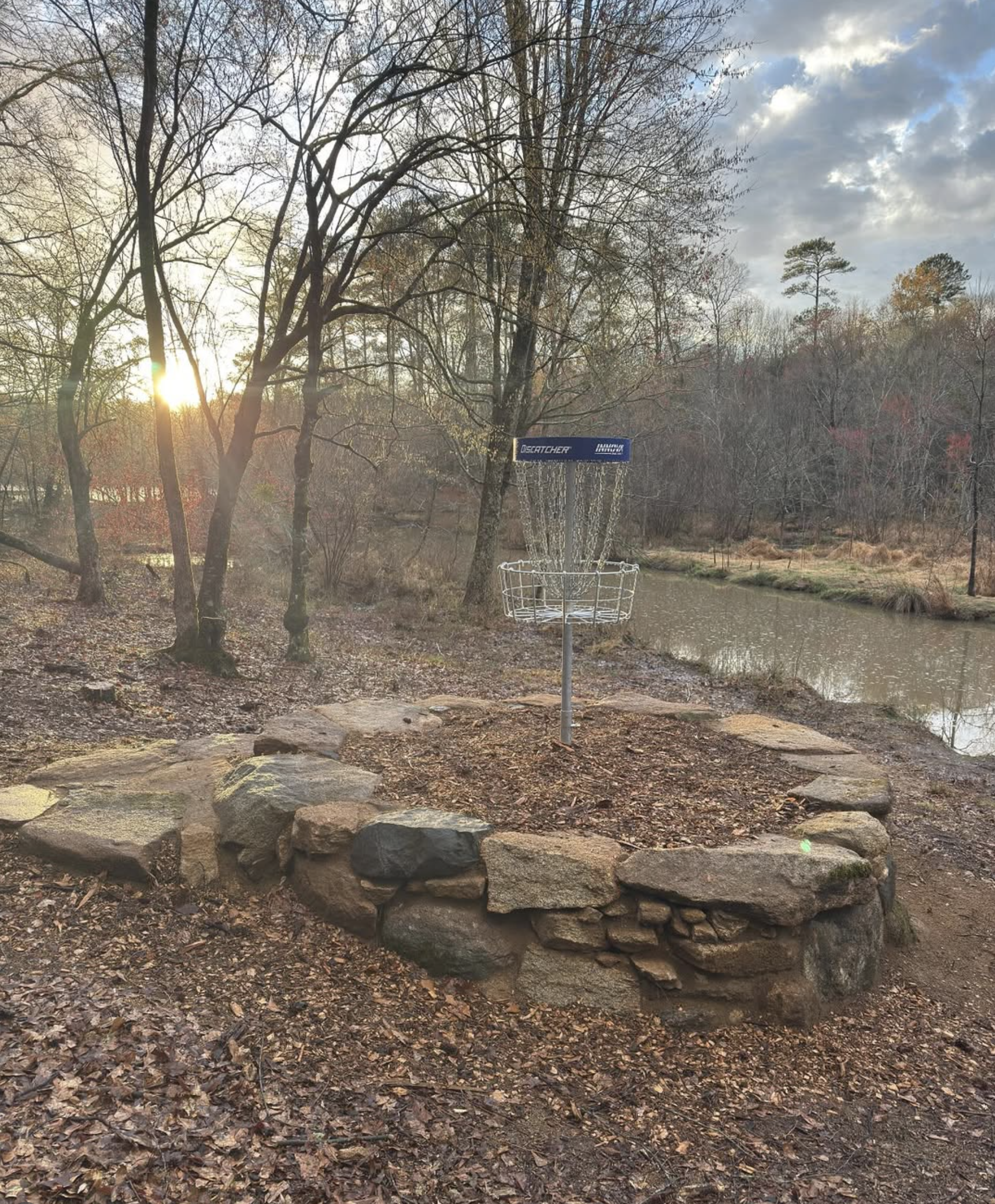 A disc golf basket on a stone circle by a river with trees and sunset in the background.