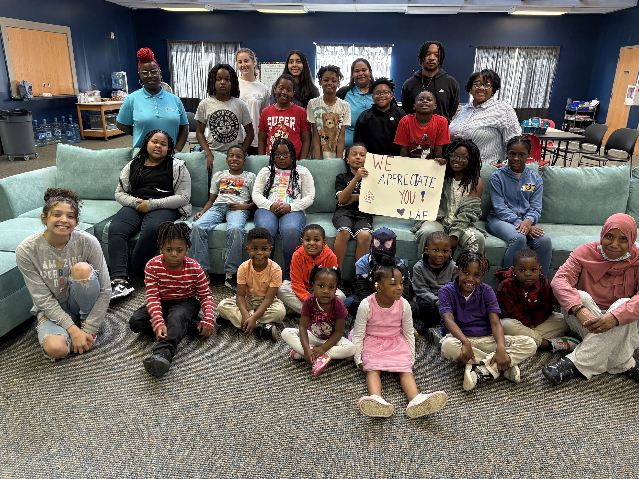 A group of children and adults gathered in a room, some seated on a turquoise couch and others sitting or kneeling on the carpeted floor, holding a sign that says, 'We appreciate you! LAE.' The room has blue walls, windows with curtains, and various items like water bottles and chairs in the background.