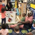 Children and an adult in a classroom or daycare setting, with educational posters and shelves in the background.