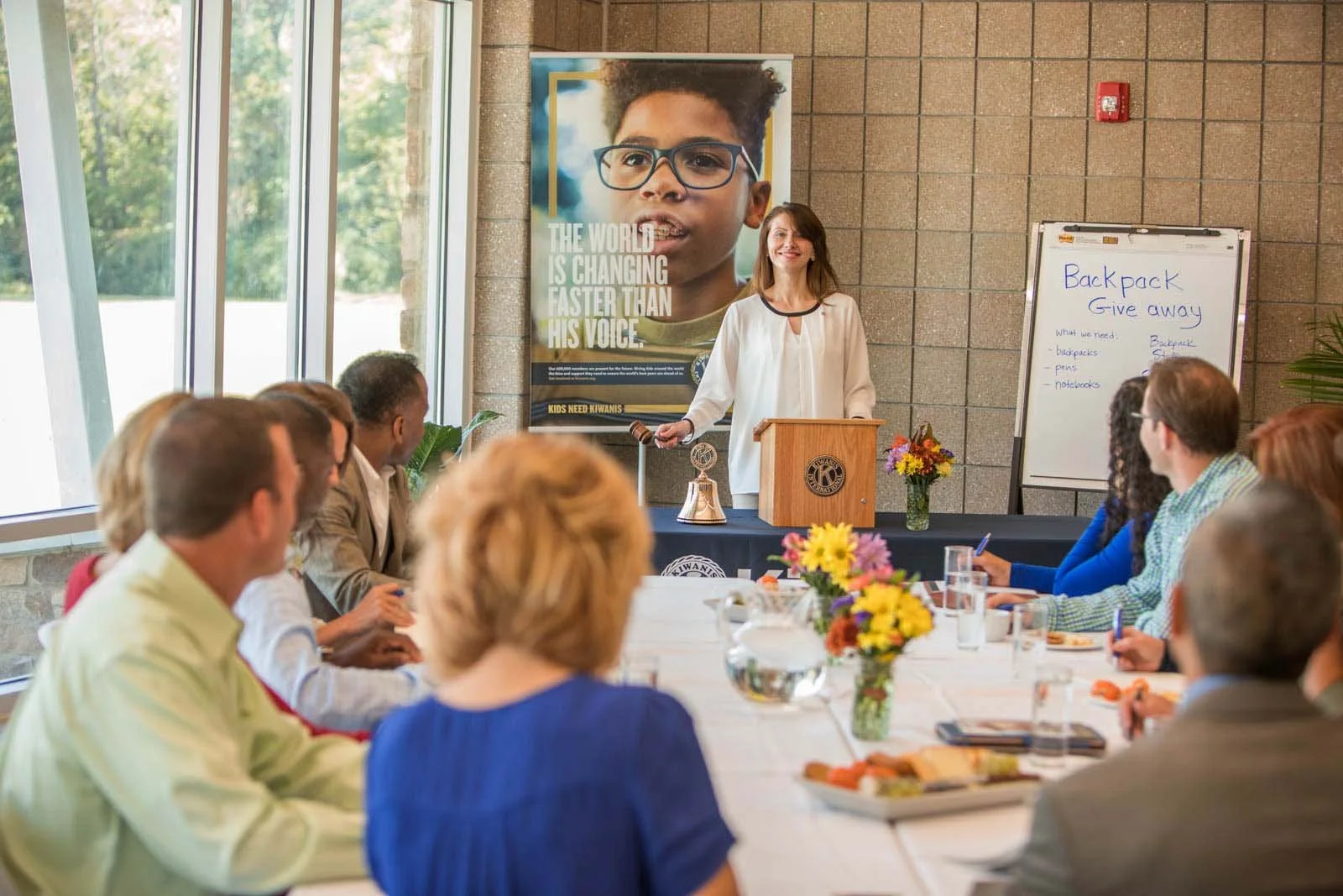 A woman giving a presentation to a group of people seated around a conference table. She is standing behind a podium with a Lions Club emblem, holding a microphone. There are notebooks, glasses of water, and flower arrangements on the table. Behind her is a large poster of a young person with glasses and text about changing the world, and a whiteboard with notes about giving away backpacks.