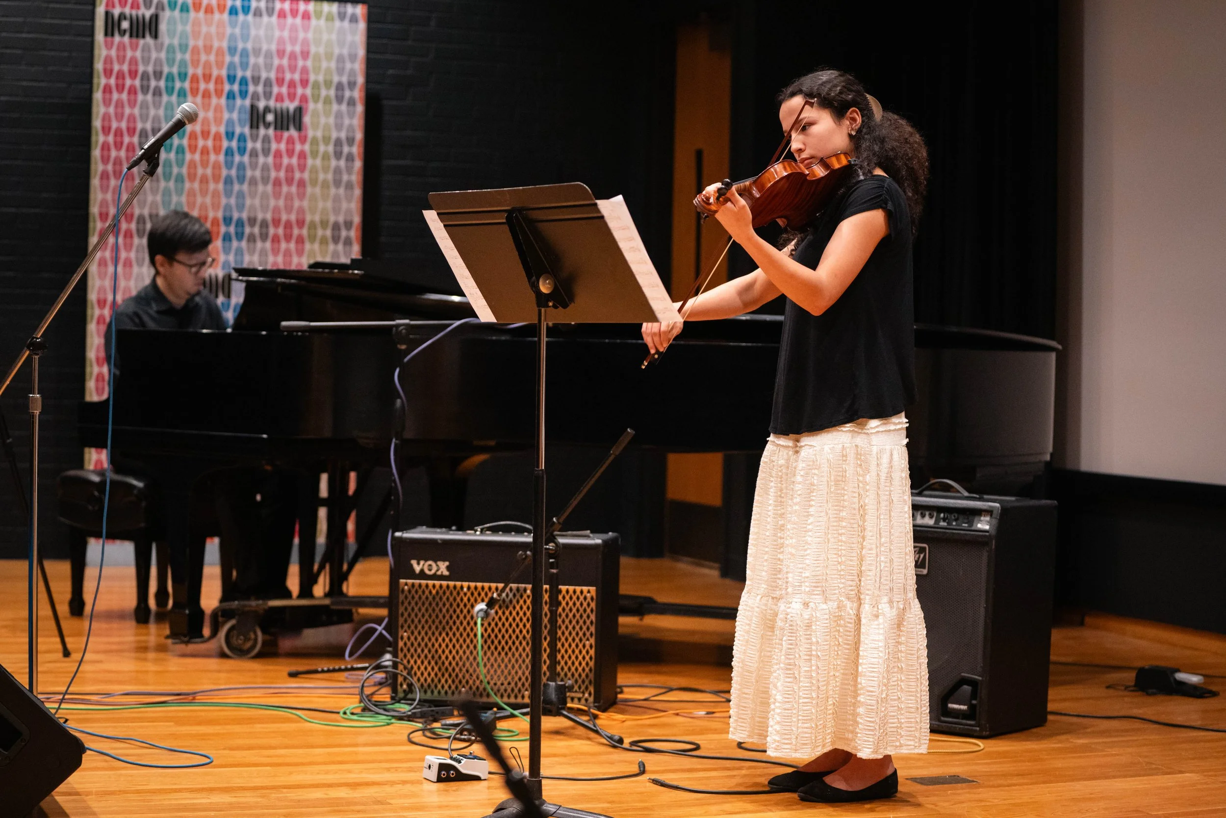 A young woman playing the violin on stage with a pianist accompanying her in the background.