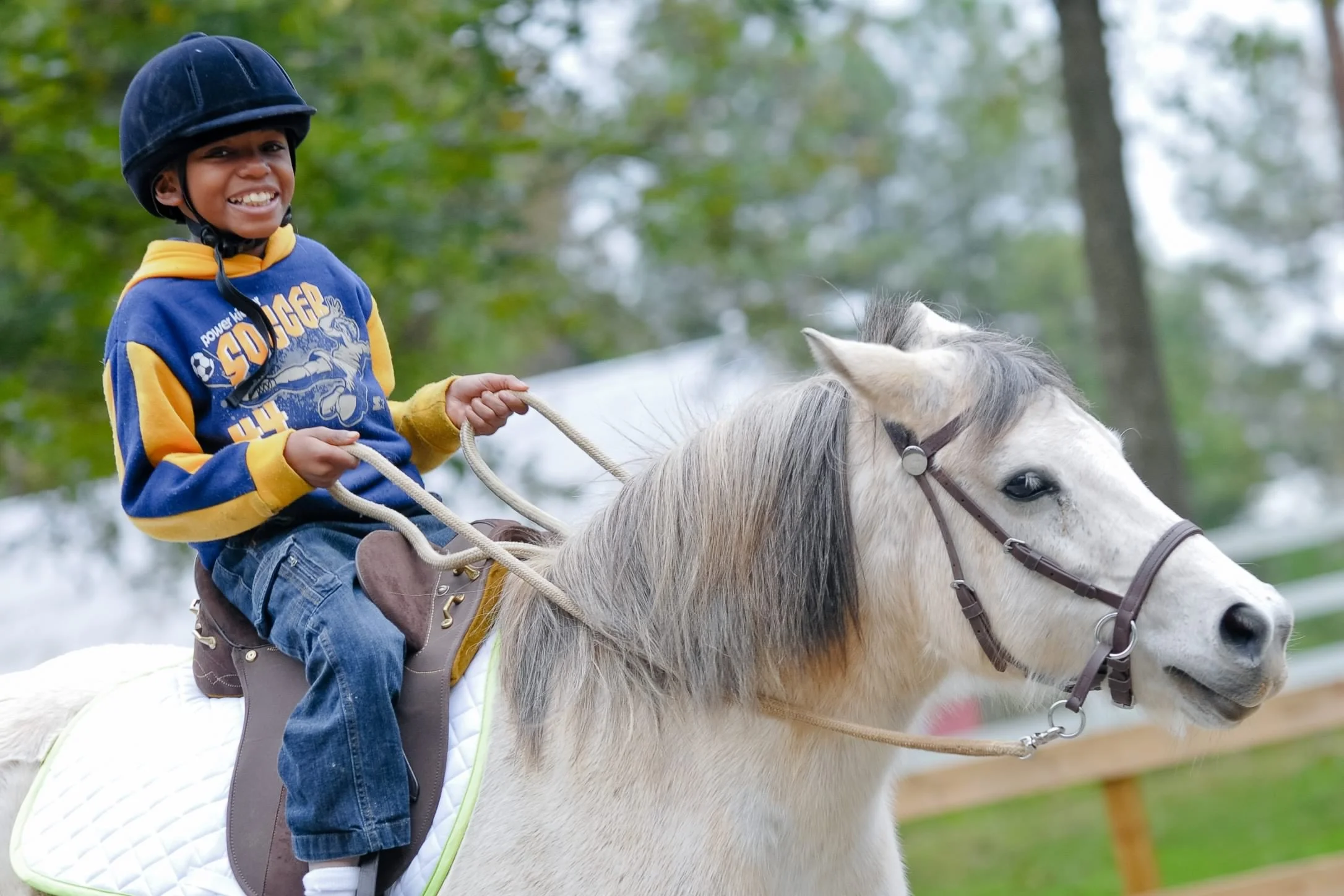A young boy smiling while riding a white horse outdoors with green trees in the background.