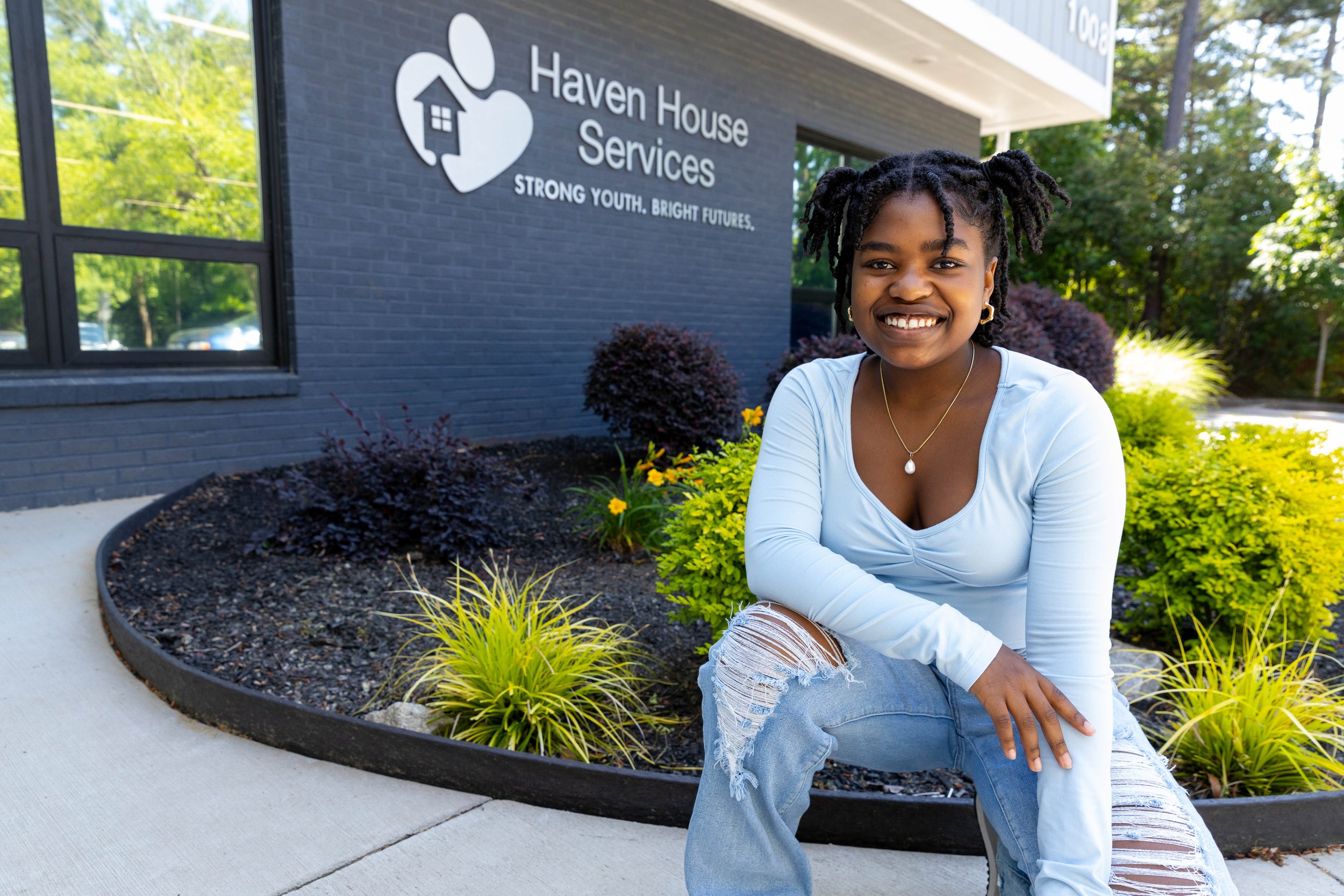 A smiling young woman with braided hair, wearing a light blue top and ripped jeans, squatting outdoors in front of a building with the sign 'Haven House Services.' The building has a dark brick exterior, and there are plants and bushes around her.