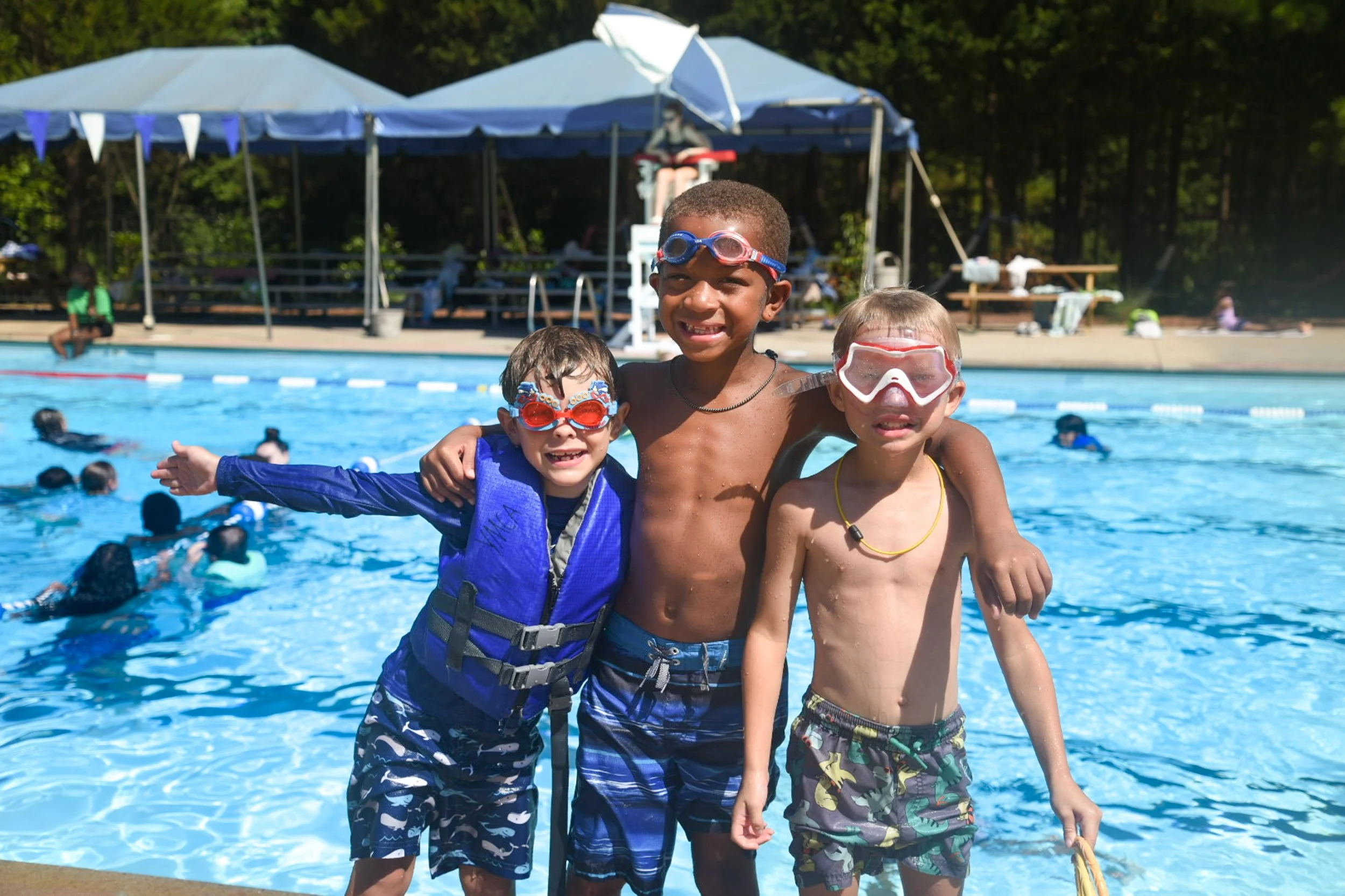 Three boys smiling and posing by the poolside at a swim party, wearing goggles and swimwear, with people swimming in the background and a large tent.