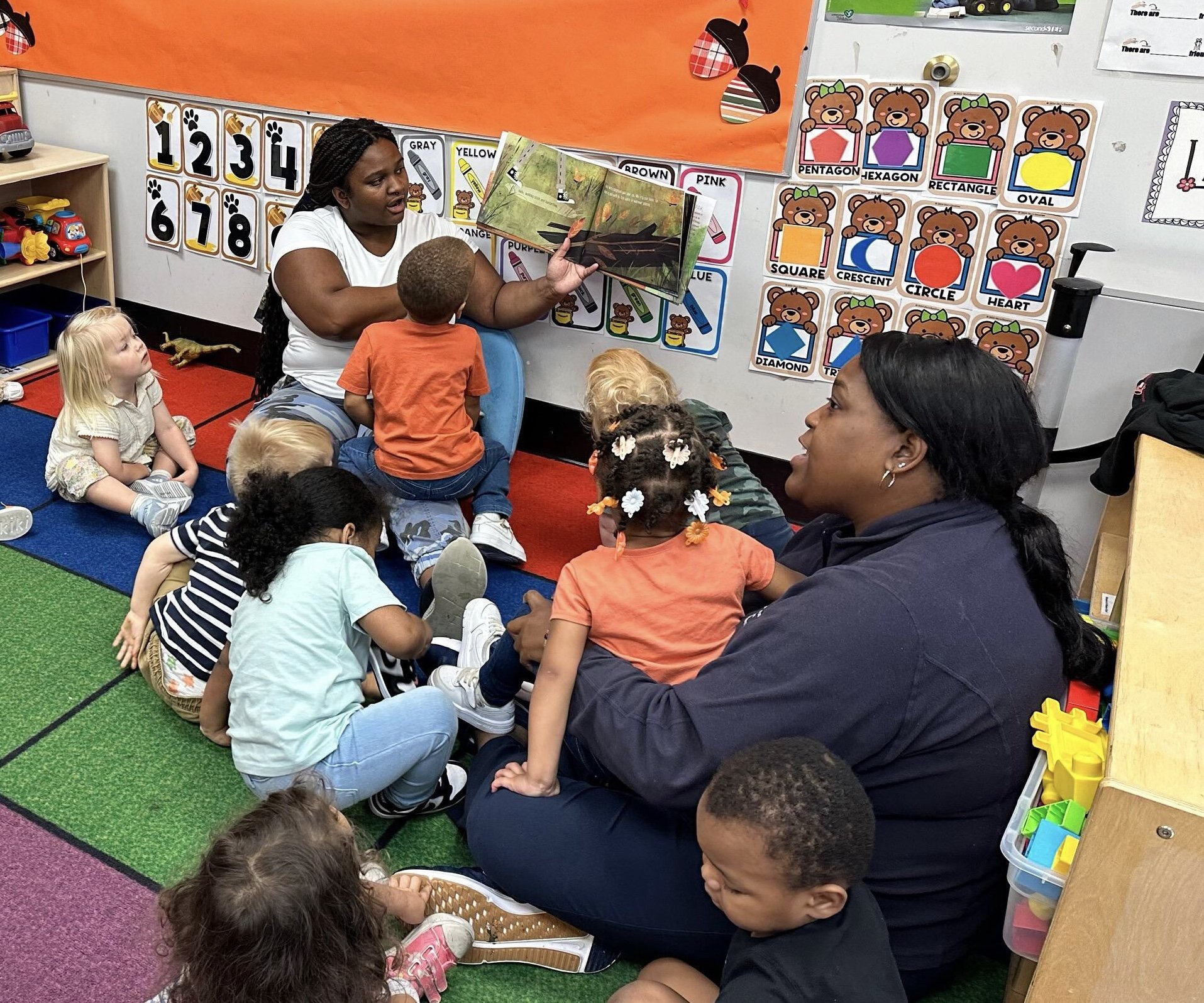 A teacher reading a book to a group of young children sitting on a colorful classroom carpet, with educational posters and toys in the background.