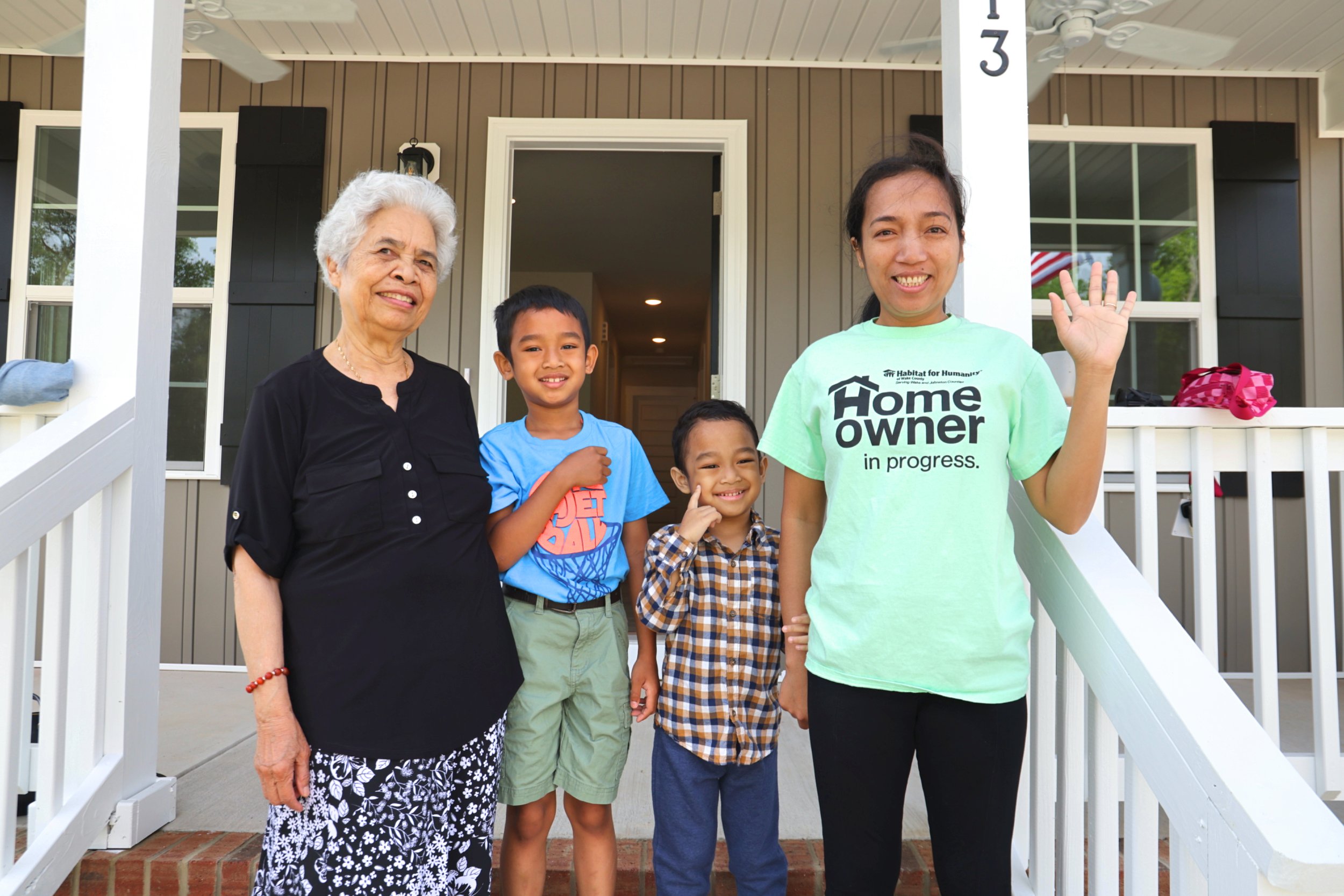 A diverse family standing on the front porch of a house, smiling and waving. The group includes an elderly woman, two young boys, a young woman wearing a 'Homeowner in progress' t-shirt, and a young girl. The house has beige siding and white railings