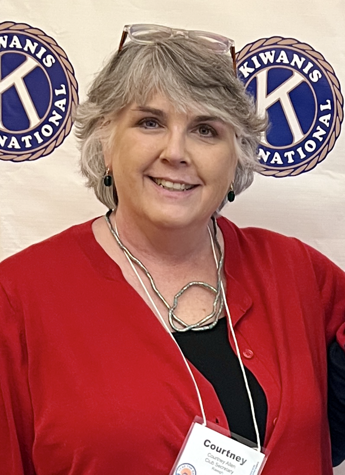 A woman with short gray hair, wearing a red blouse, glasses on her head, and a black top underneath. She has green earrings and a silver necklace. She is smiling and standing in front of a white backdrop with two blue and gold logos that say 'Kiwanis International'.