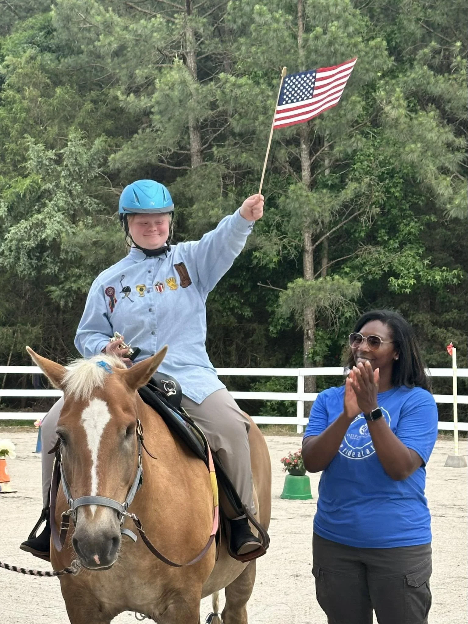 A boy in a blue helmet and gray shirt sitting on a brown horse, holding an American flag, with a woman clapping nearby in a sandy outdoor area surrounded by green trees.