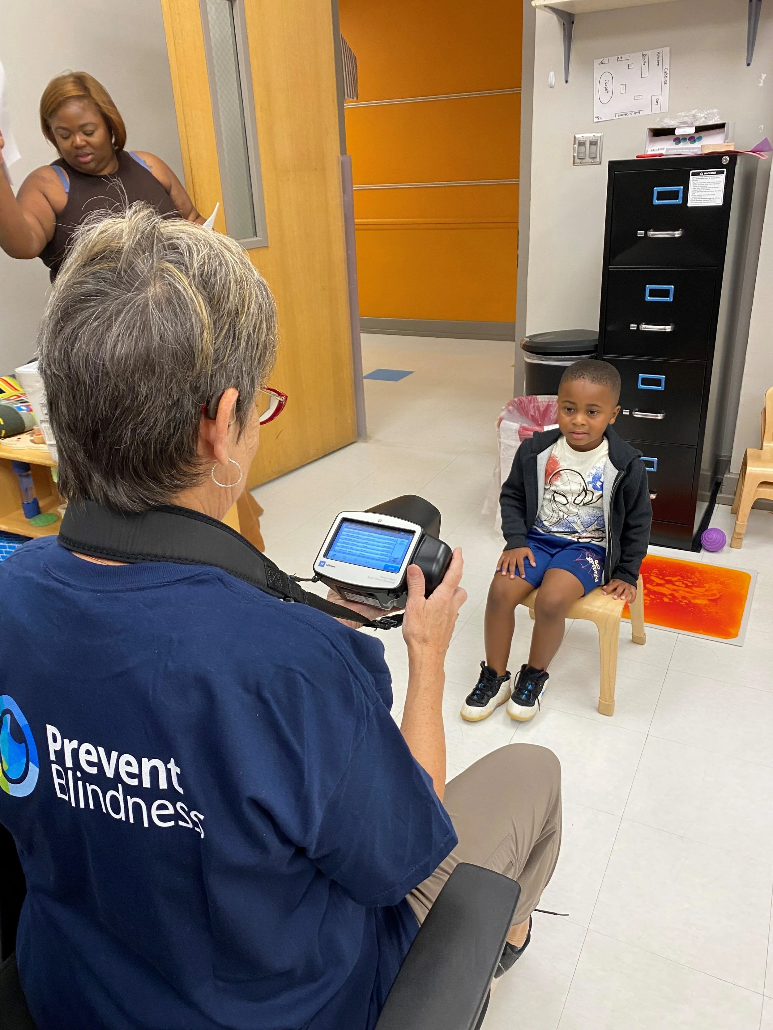 A woman with short gray hair and glasses, wearing a navy blue shirt with the words 'Prevent Blindness' on the back, is seated while performing a vision test on a young boy with short hair, wearing a Spider-Man T-shirt, a black jacket, and blue shorts