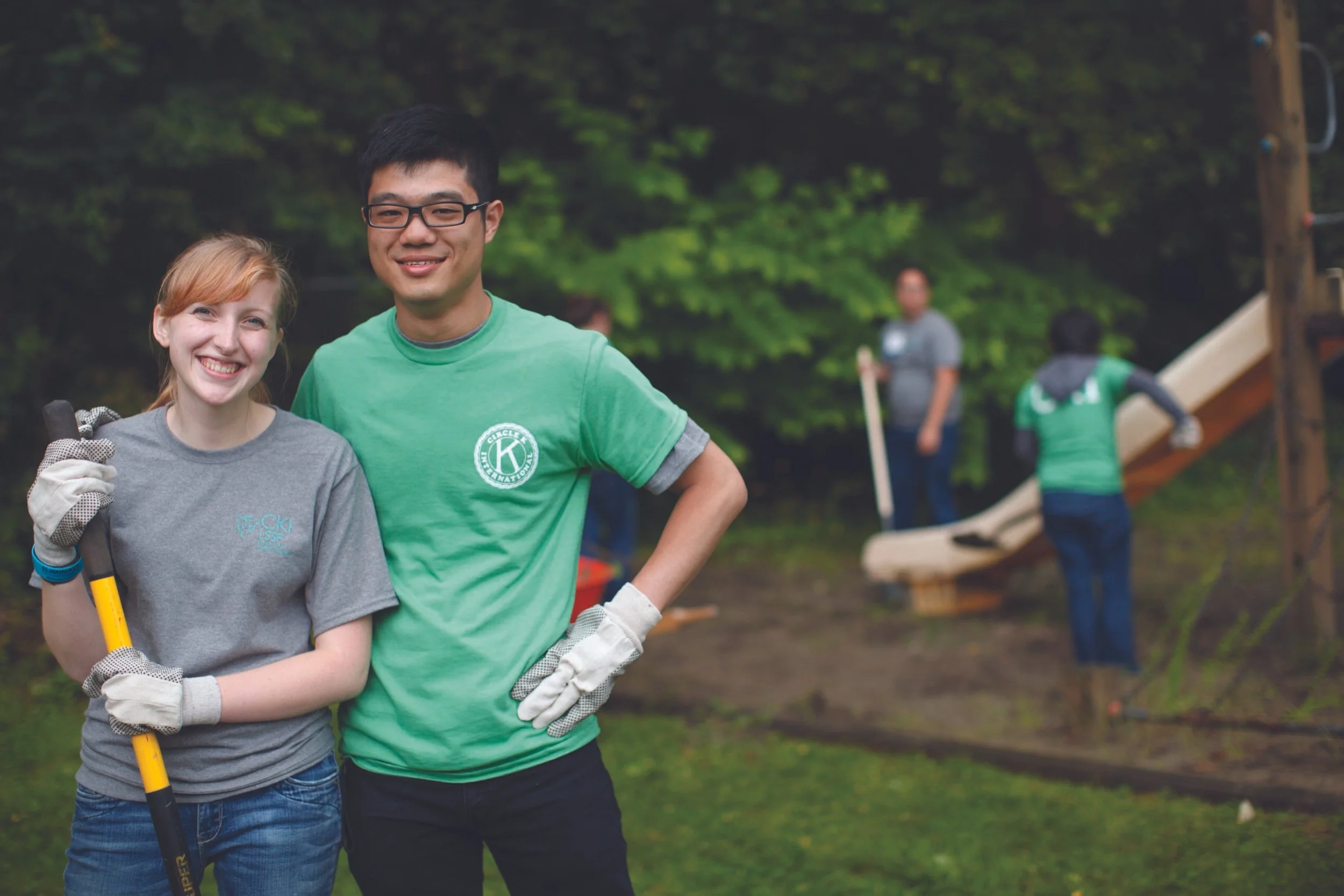 Two people smiling at the camera, one woman and one man, standing outdoors in a green area, wearing gloves and holding a tool. In the background, three other individuals are working on a playground structure, with trees surrounding the area.