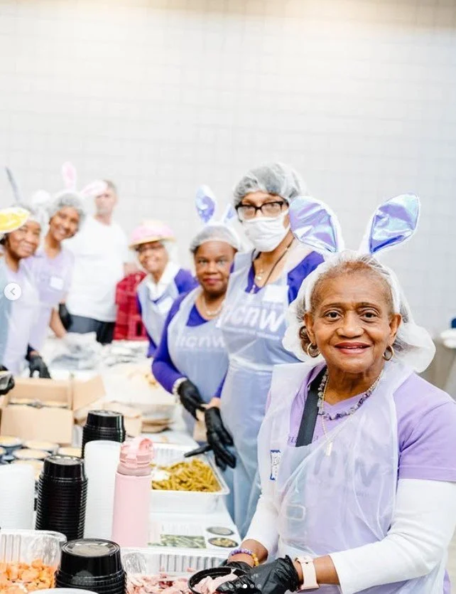 Senior woman wearing bunny ears, purple shirt, purple apron, and black gloves smiling at a community meal event with volunteers in the background.