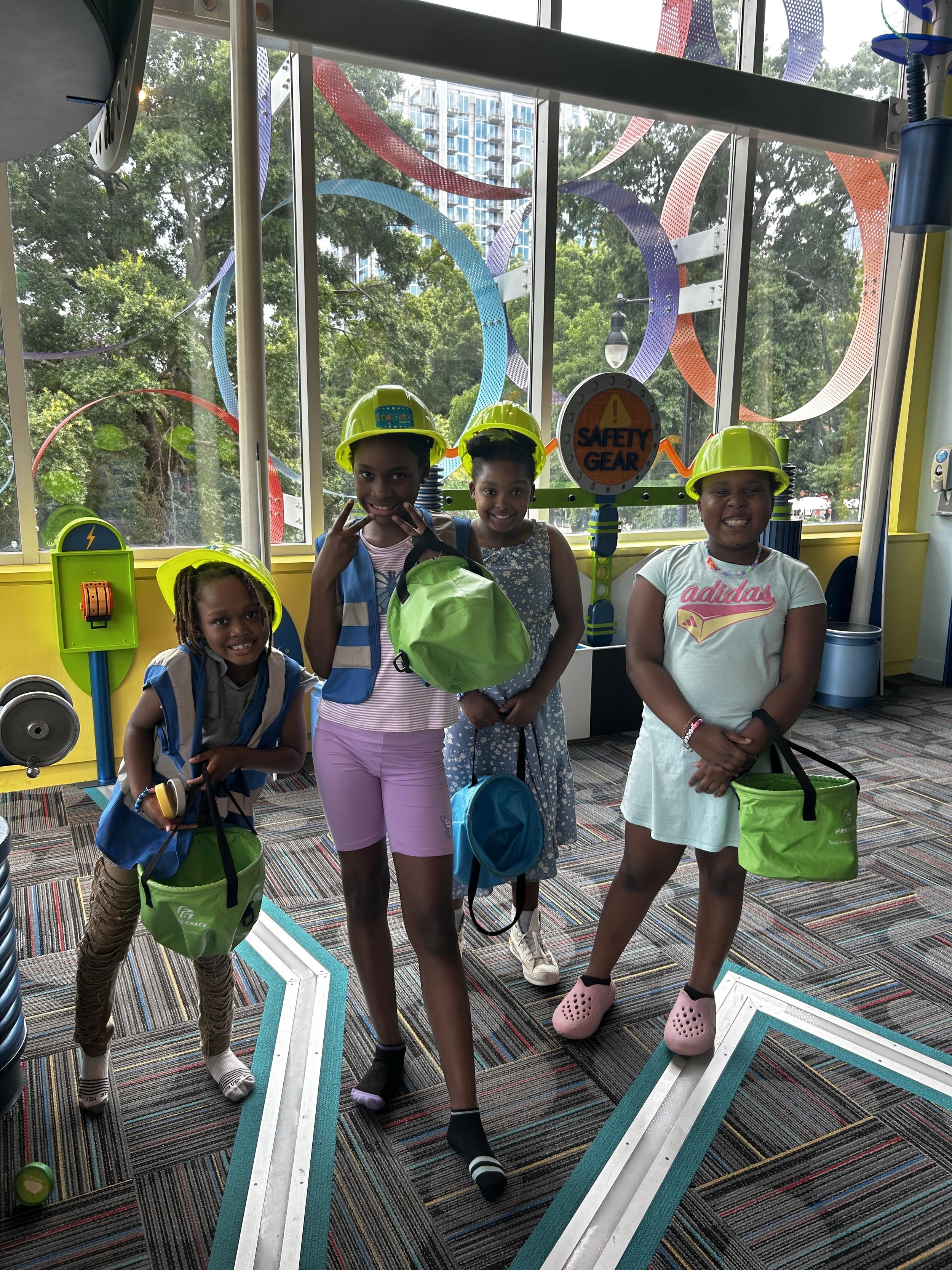 Four children wearing safety helmets and carrying bags inside an educational play area with colorful decor and large windows showing trees and a building outside.