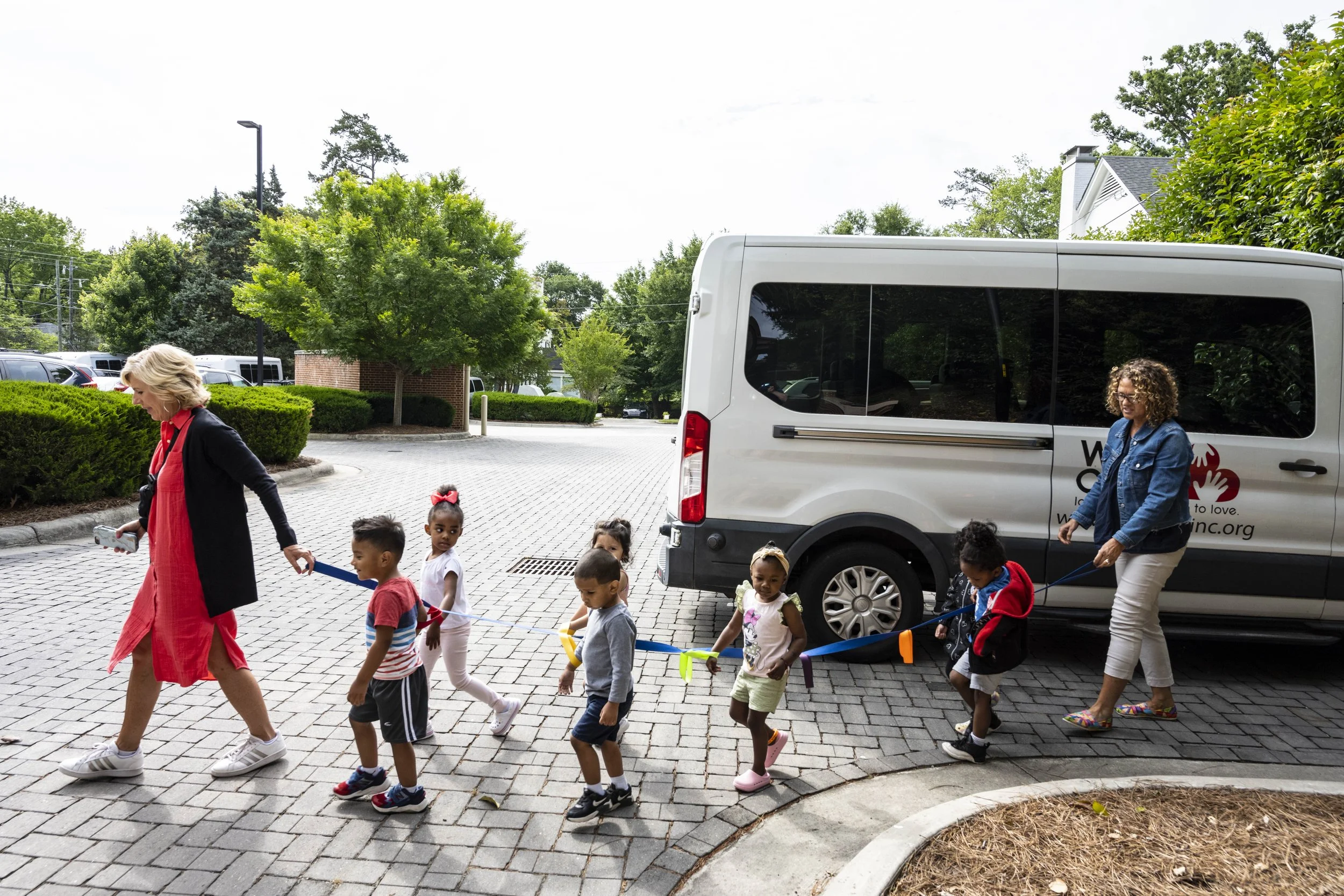 A woman leading a line of children on a walk outside near a van parked on a paved area surrounded by trees and greenery.