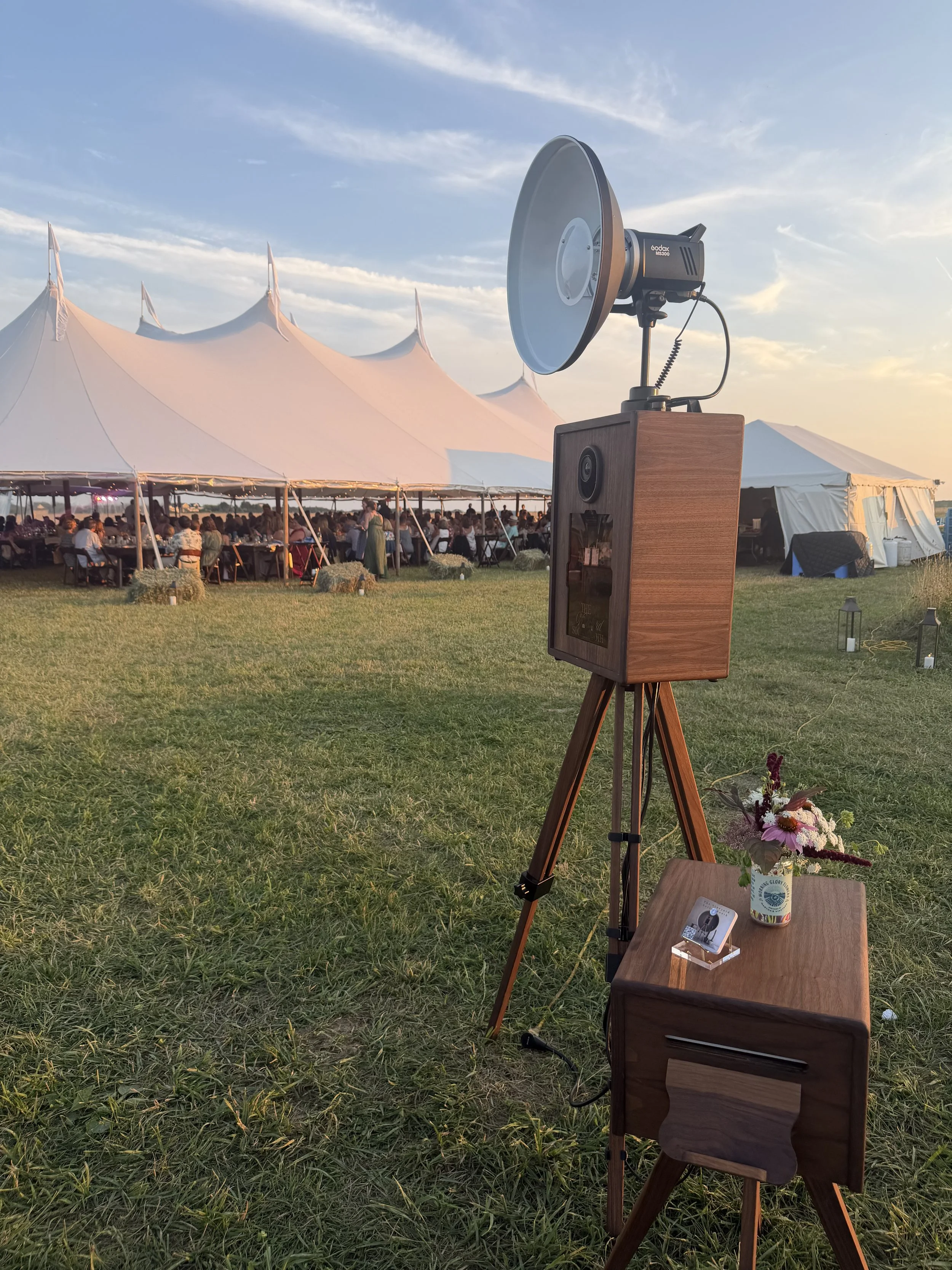 Outdoor event under large white tent with guests seated at tables, our handcrafted wooden photo booth, and a small table with a flower arrangement.