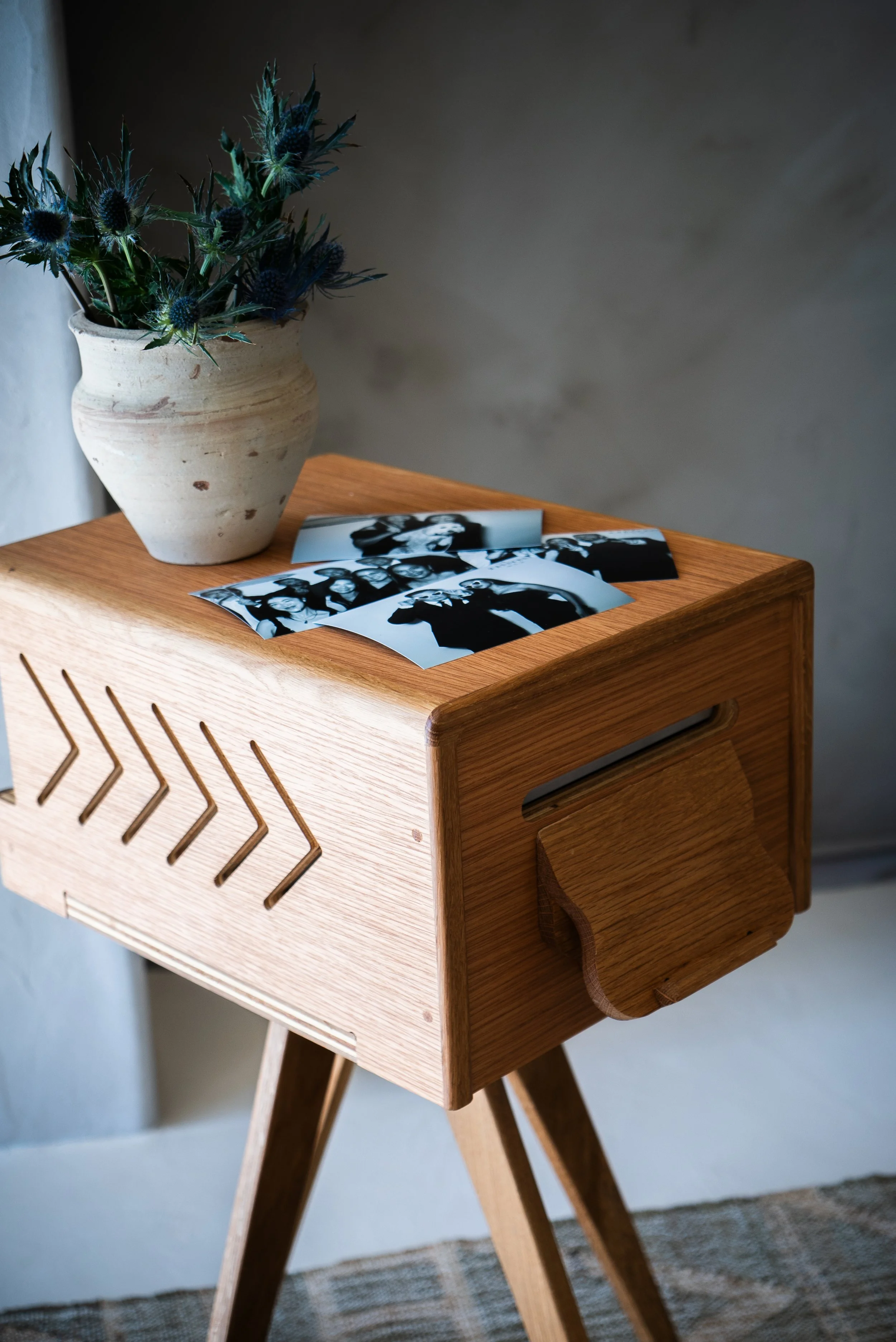 A wooden side table with three black and white photographs and a white vase with blue thistle flowers on top, set against a light gray wall.