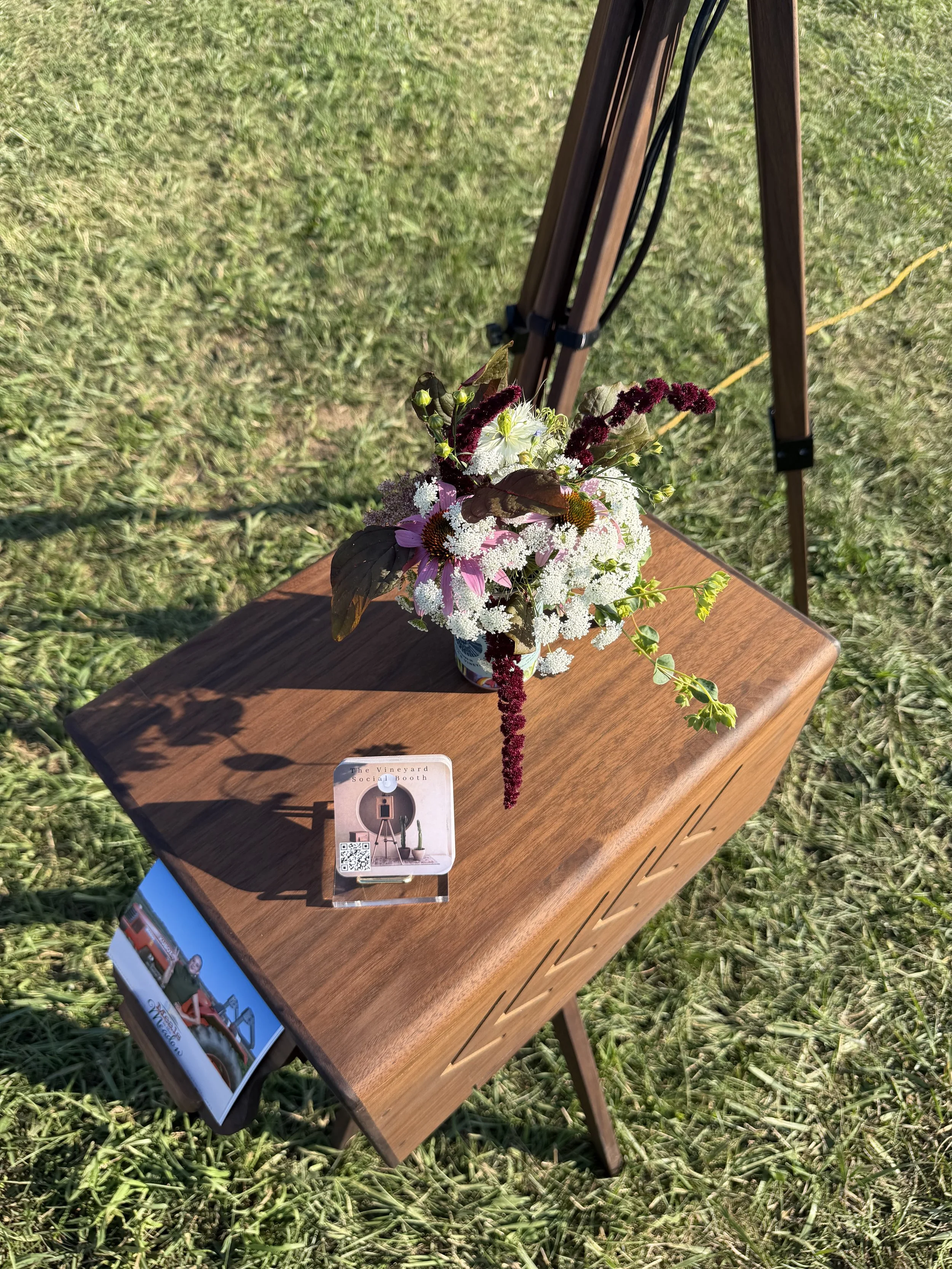 A floral arrangement of white, purple, and dark red flowers on a wooden table outdoors, with a sign and a brochure underneath.