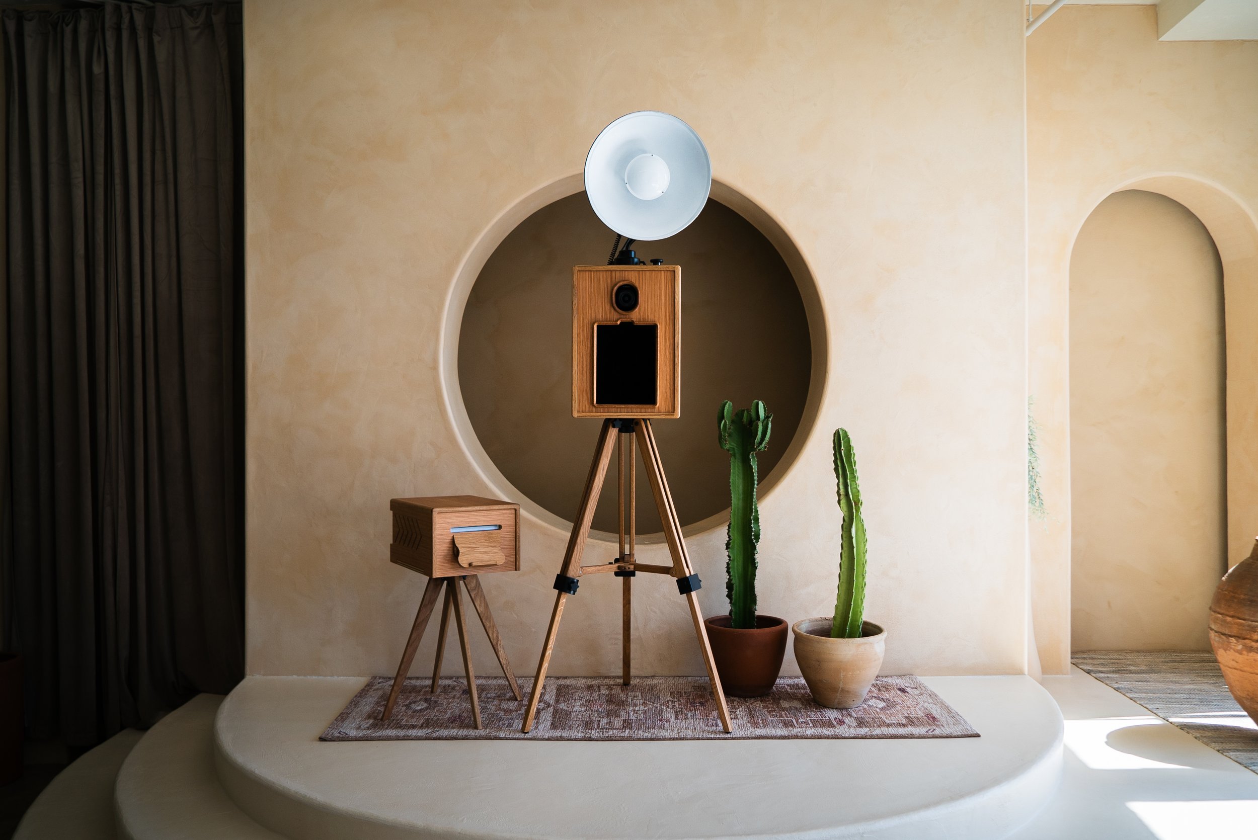 A vintage wooden speaker on a tripod stand, with an old-fashioned spotlight or studio light mounted on top, placed on a small area rug in front of a beige wall with a circular cutout. Next to the speaker are two potted cactus plants, one in a brown pot and the other in a light-colored pot. On the left side, there is a small wooden side table with a drawer.