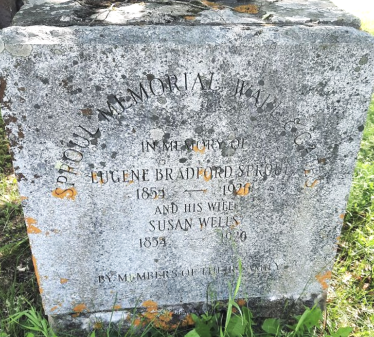 A weathered stone surrounded by grass and some orange lichen, in dedication to the Sprouls.