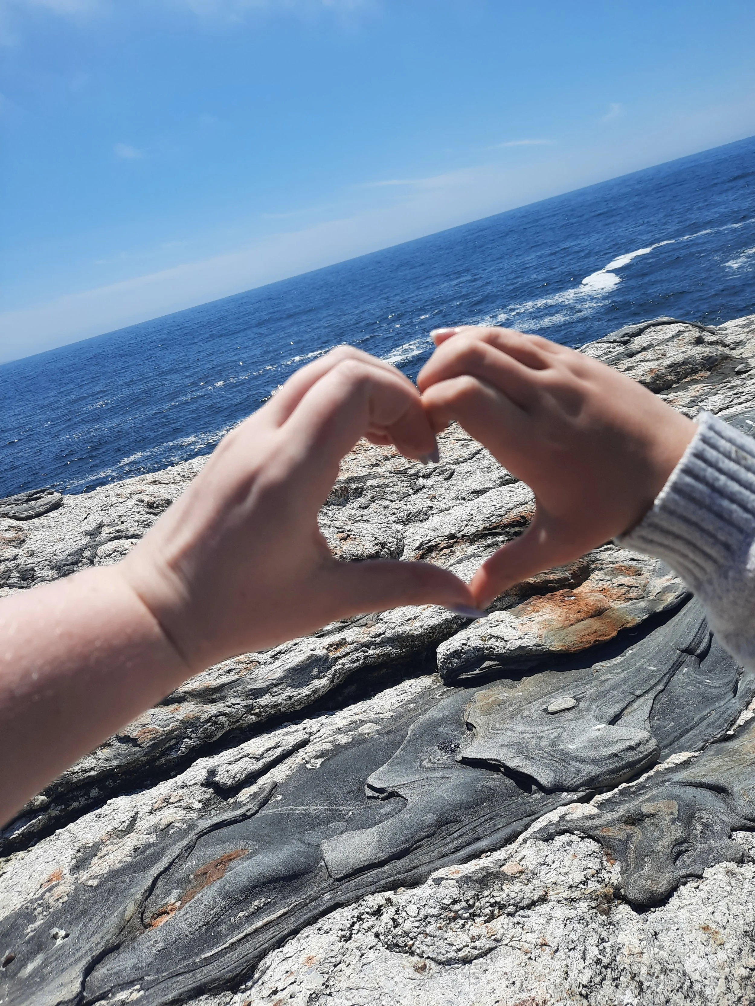 Two hands forming a heart shape in front of a rocky coastline and ocean under a blue sky.