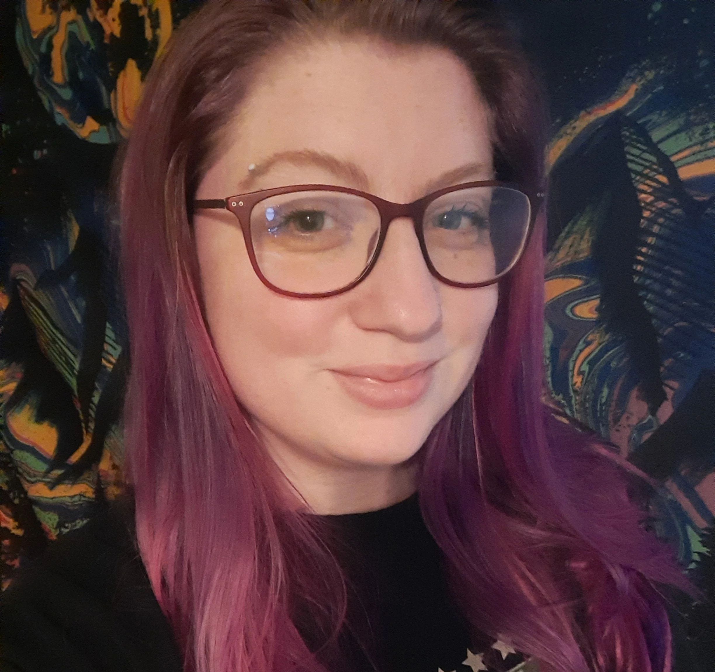 Close-up of a woman with pink and purple hair, wearing glasses, standing against a colorful patterned fabric background.