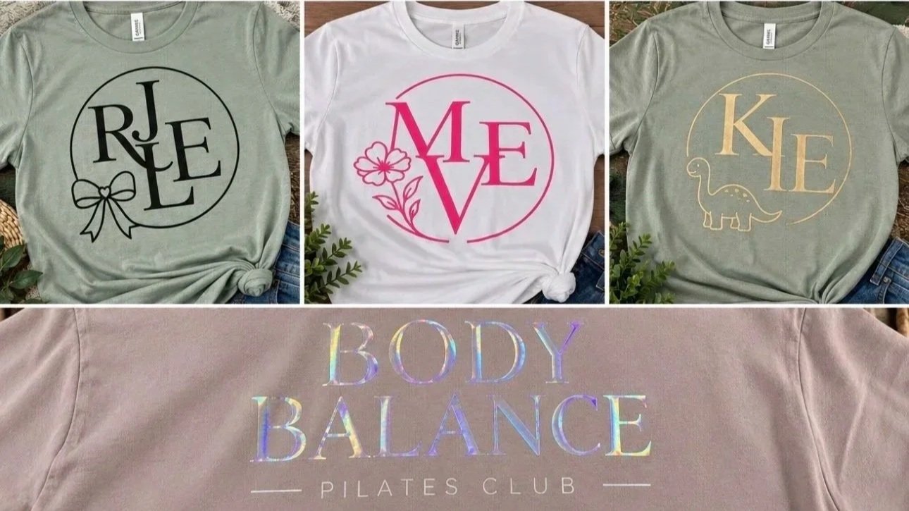 Four t-shirts with different designs and texts, laid out on a flat surface. The first shirt is light gray with the word "RELEASE" inside a circle, decorated with a bow. The second shirt is white with the word "LOVE" inside a circle, accented with a flower and leaves. The third shirt is light green with the word "KIND" inside a circle, featuring a small dinosaur illustration. The fourth shirt is light pink with holographic text reading "BODY BALANCE PILATES CLUB."