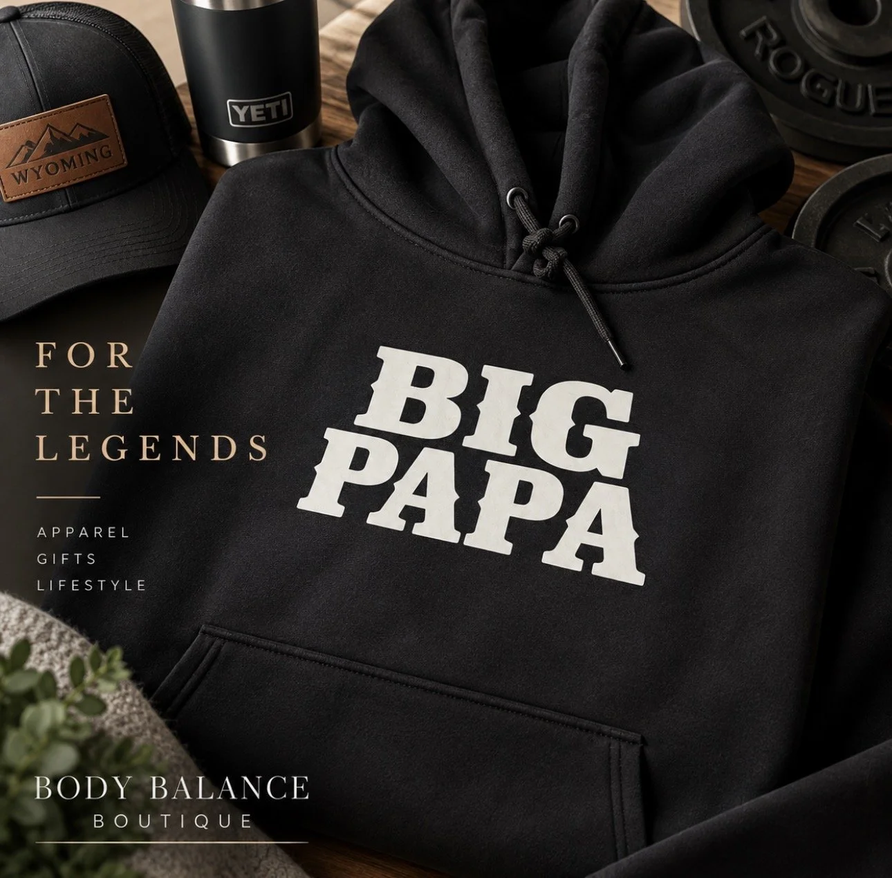 Black hoodie with white text reading 'BIG PAPA' surrounded by fitness equipment including a dumbbell and a tumbler, as well as a branded baseball cap in the background.