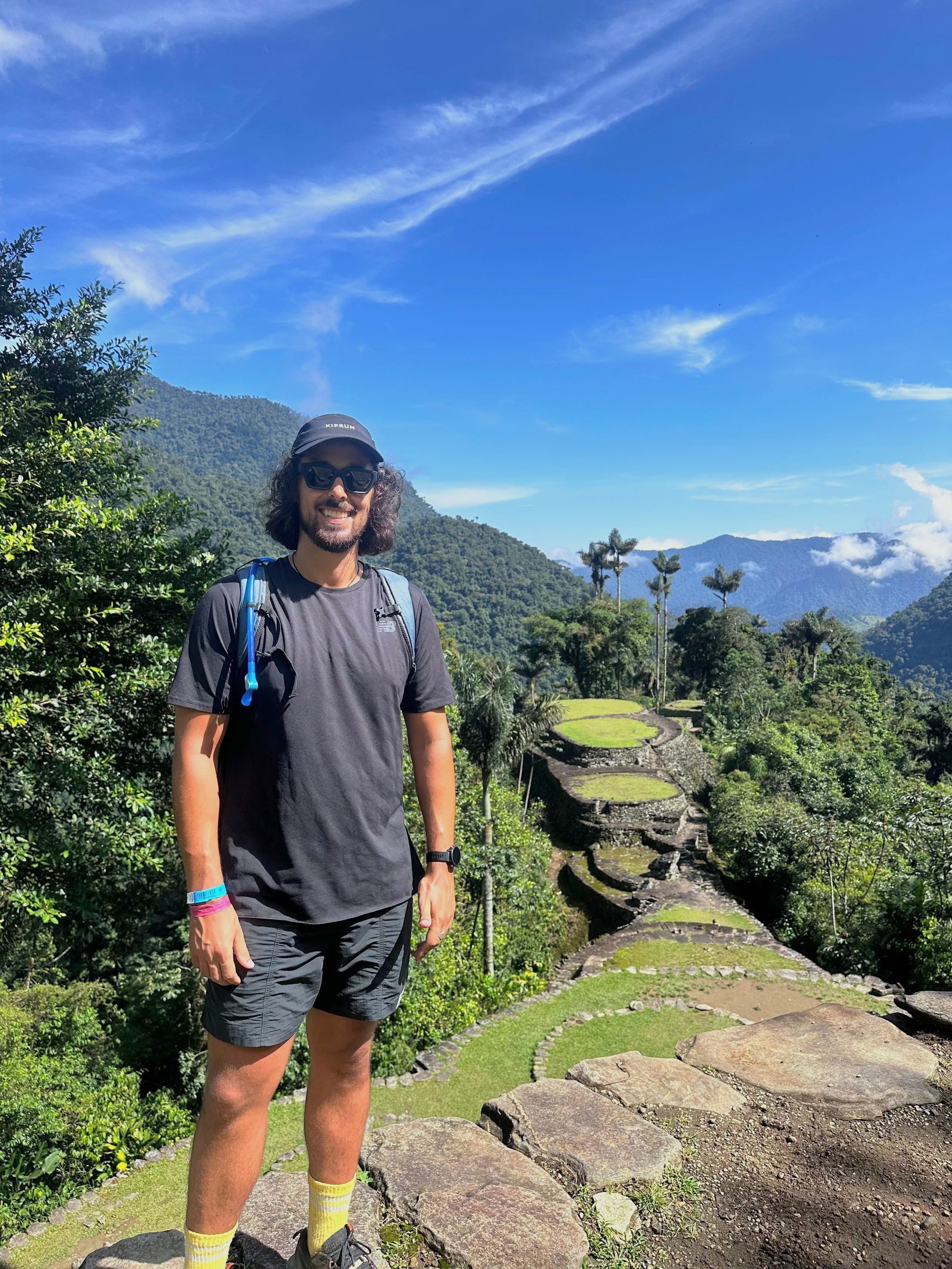 Man standing on ancient stone ruins in lush green mountains with clear blue sky overhead.