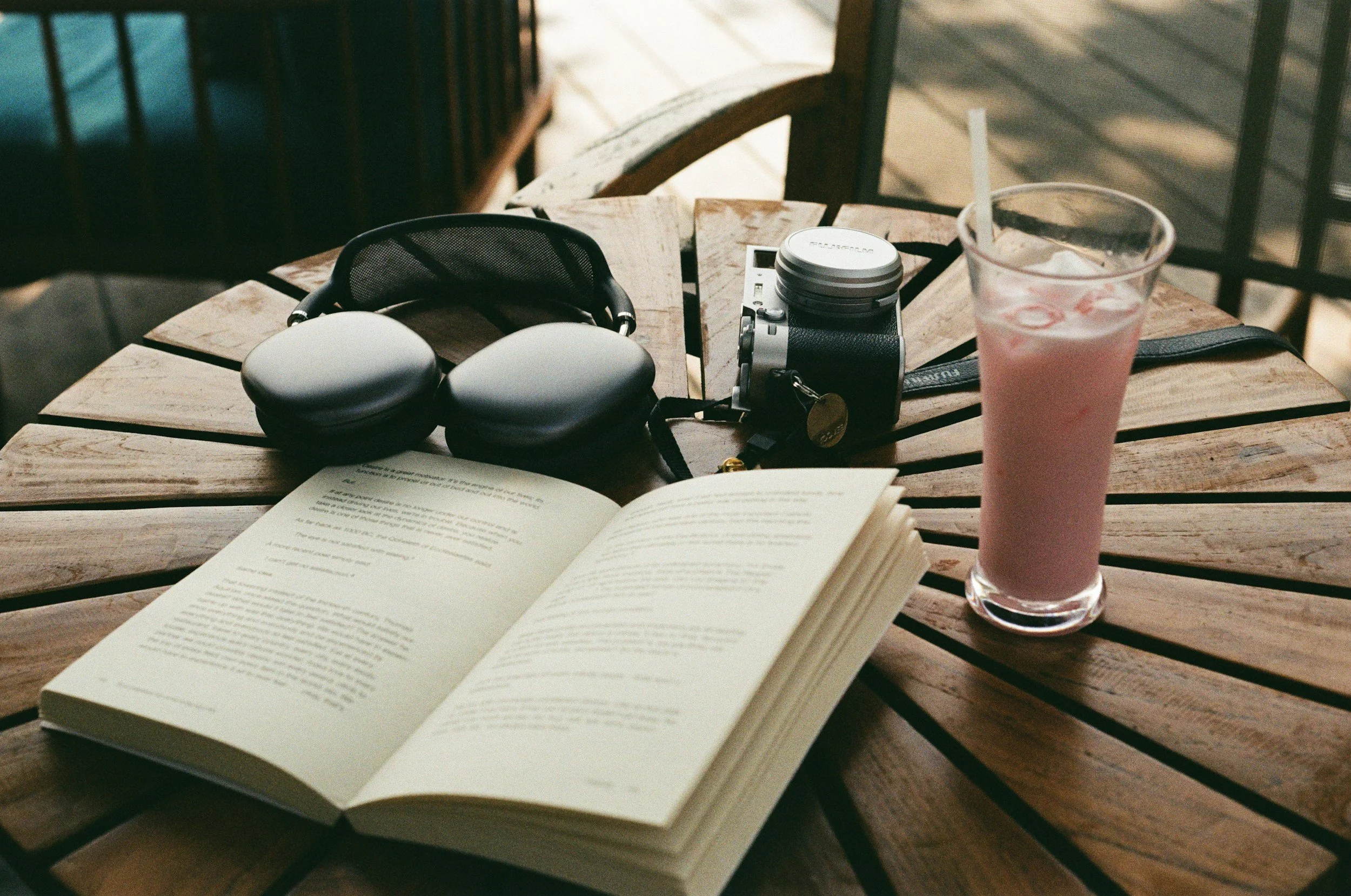A wooden outdoor table with an open book, a pair of black over-ear headphones, a camera, a drink with a straw, and a black wallet on it.