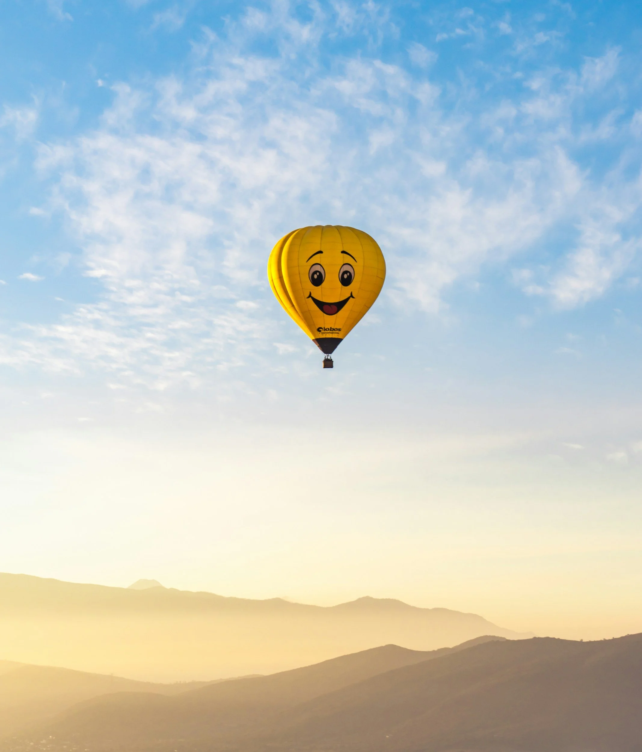 Yellow hot air balloon with a smiling emoji face flying in a blue sky with clouds and mountain silhouettes in the background.