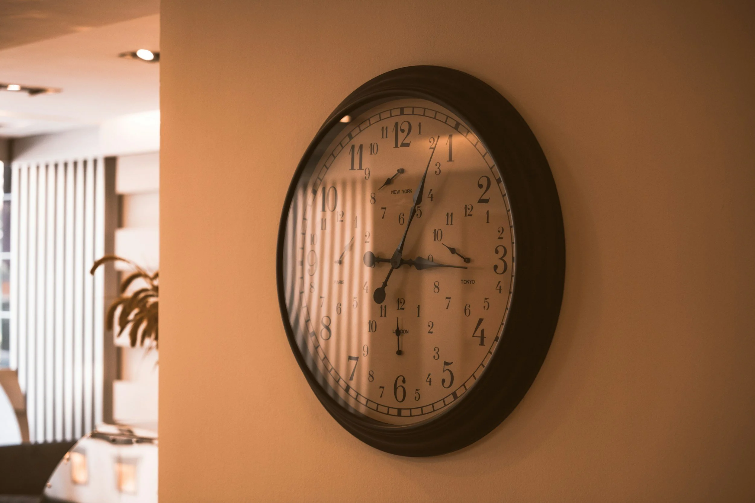 A large round wall clock showing approximately 3:02, with a black frame and a white face, mounted on a beige wall in an indoor setting with blurred background.