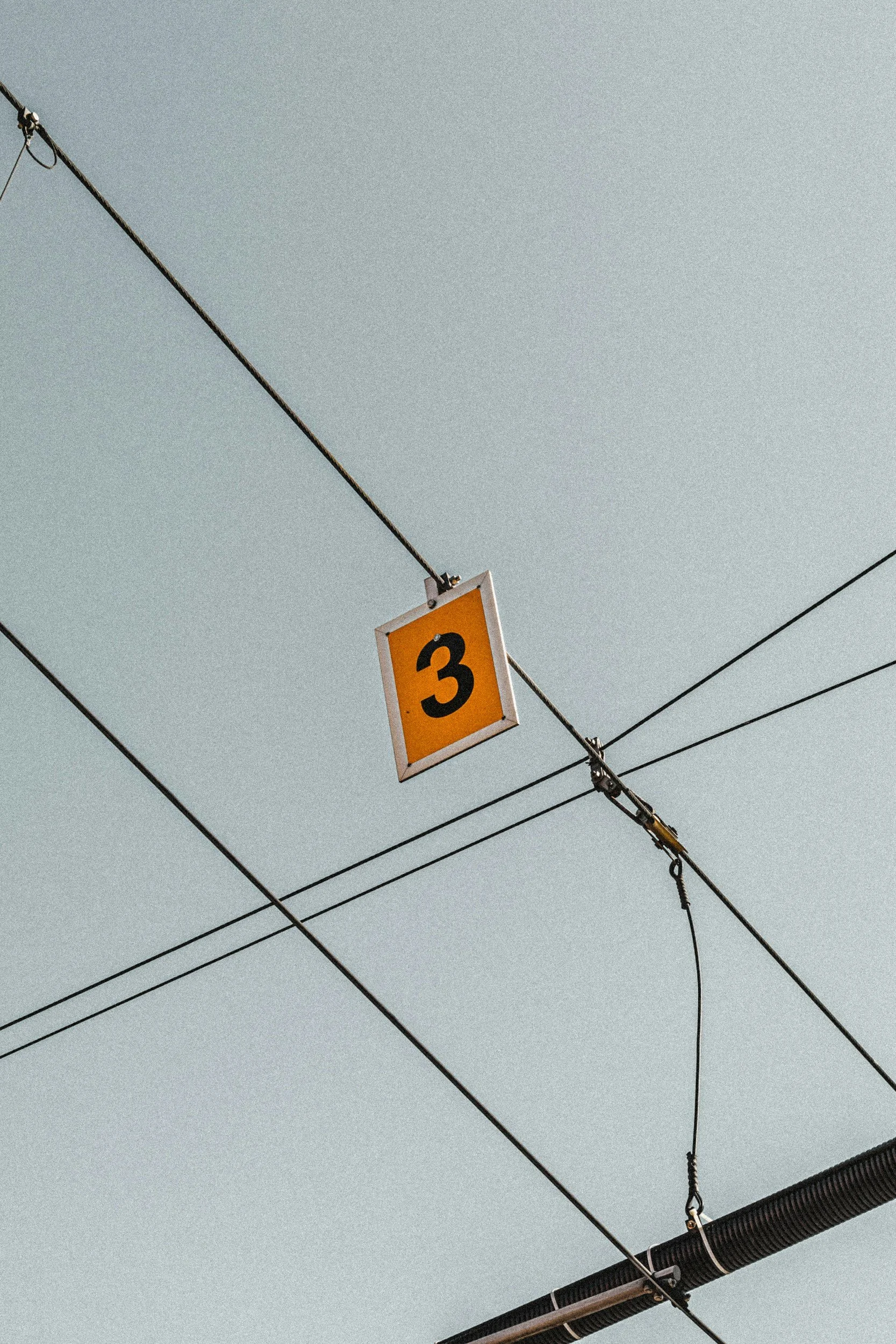 A yellow sign with a black number 3, hanging among overhead wires and cables against a clear sky.