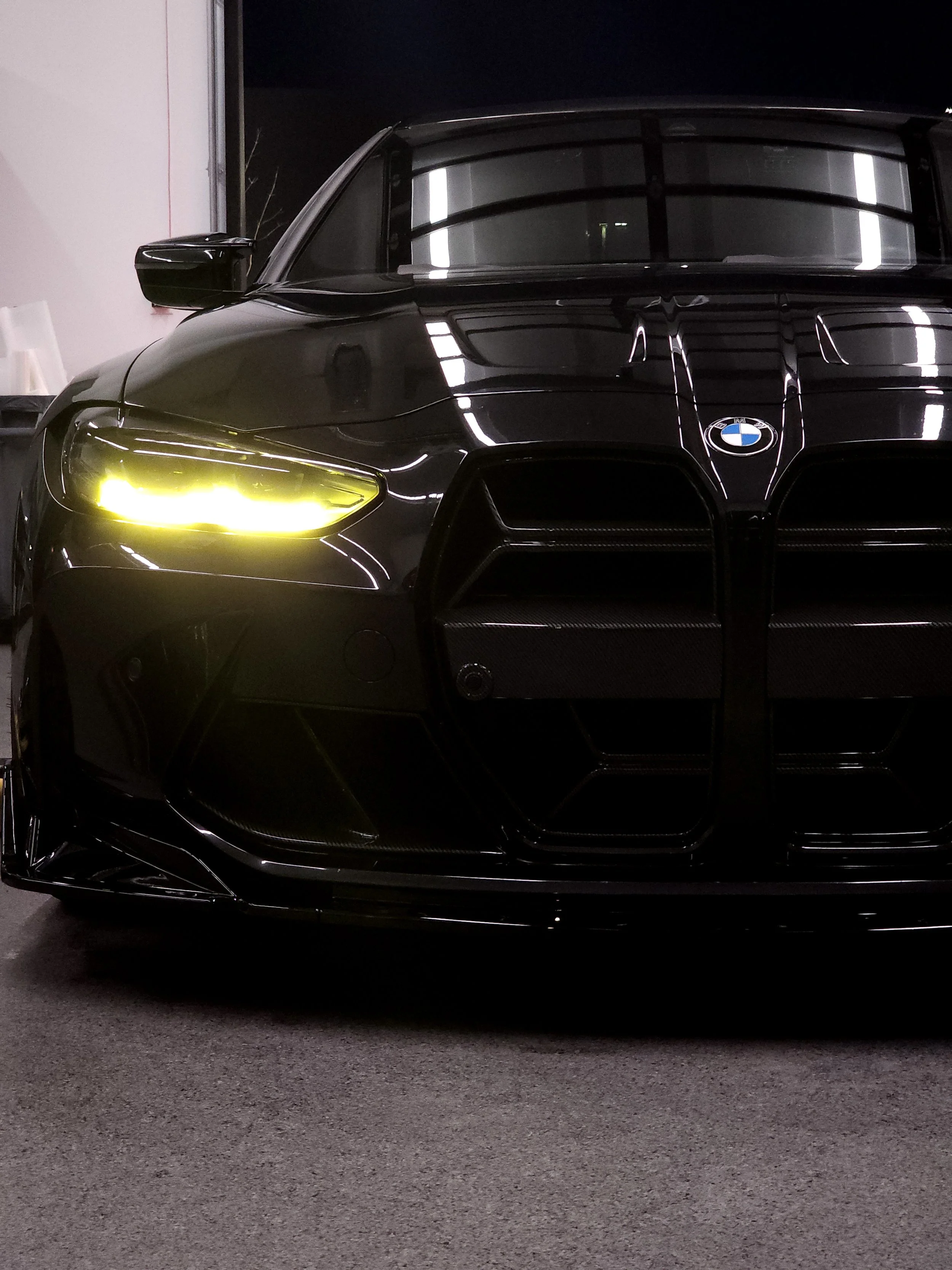 A black BMW car parked in a garage at night, with reflections of fluorescent lights on the car's surface.