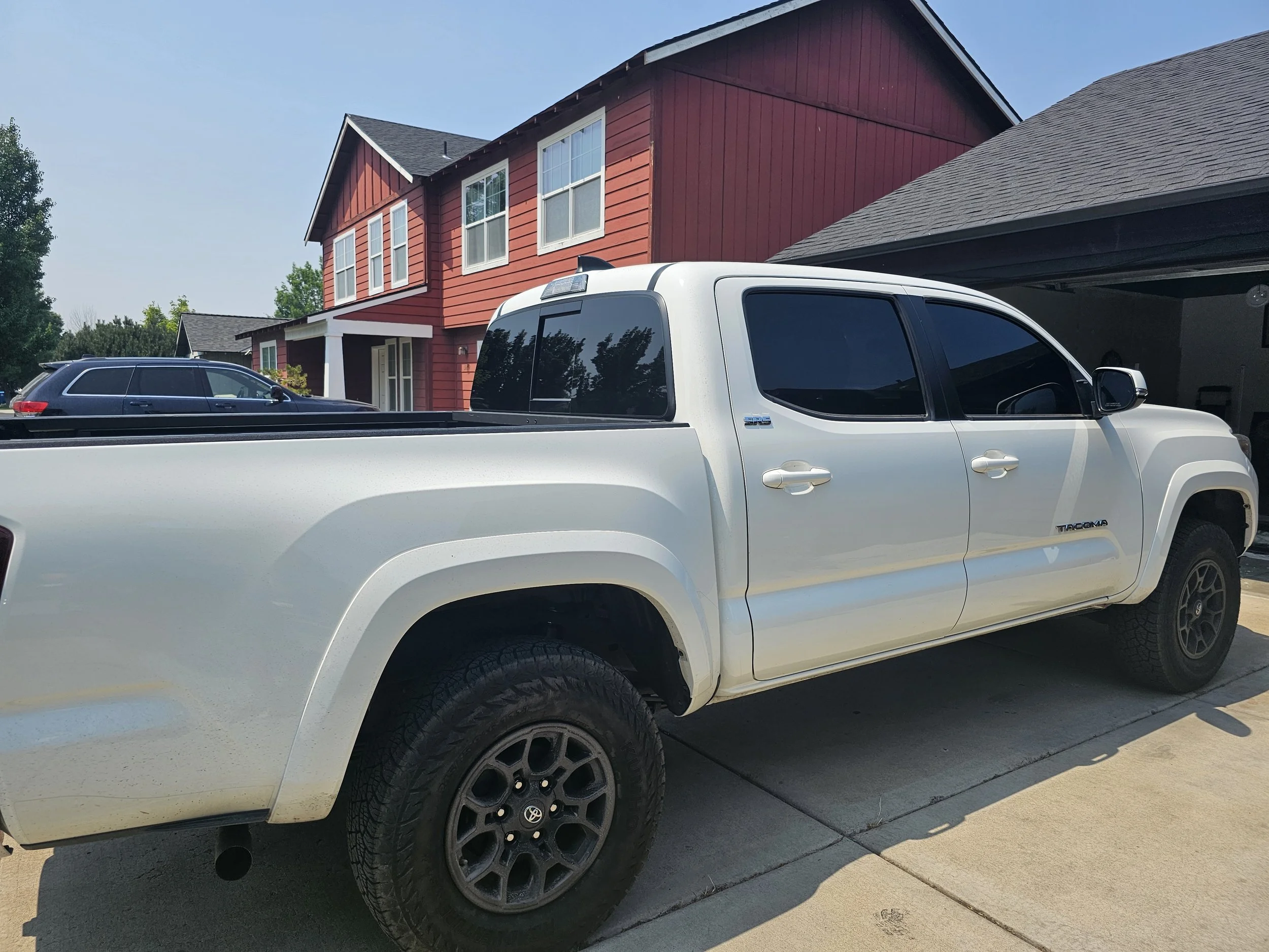 A white Toyota Tacoma pickup truck parked on the driveway in front of a red, two-story house with multiple windows. The house has a dark roof and the driveway is concrete.