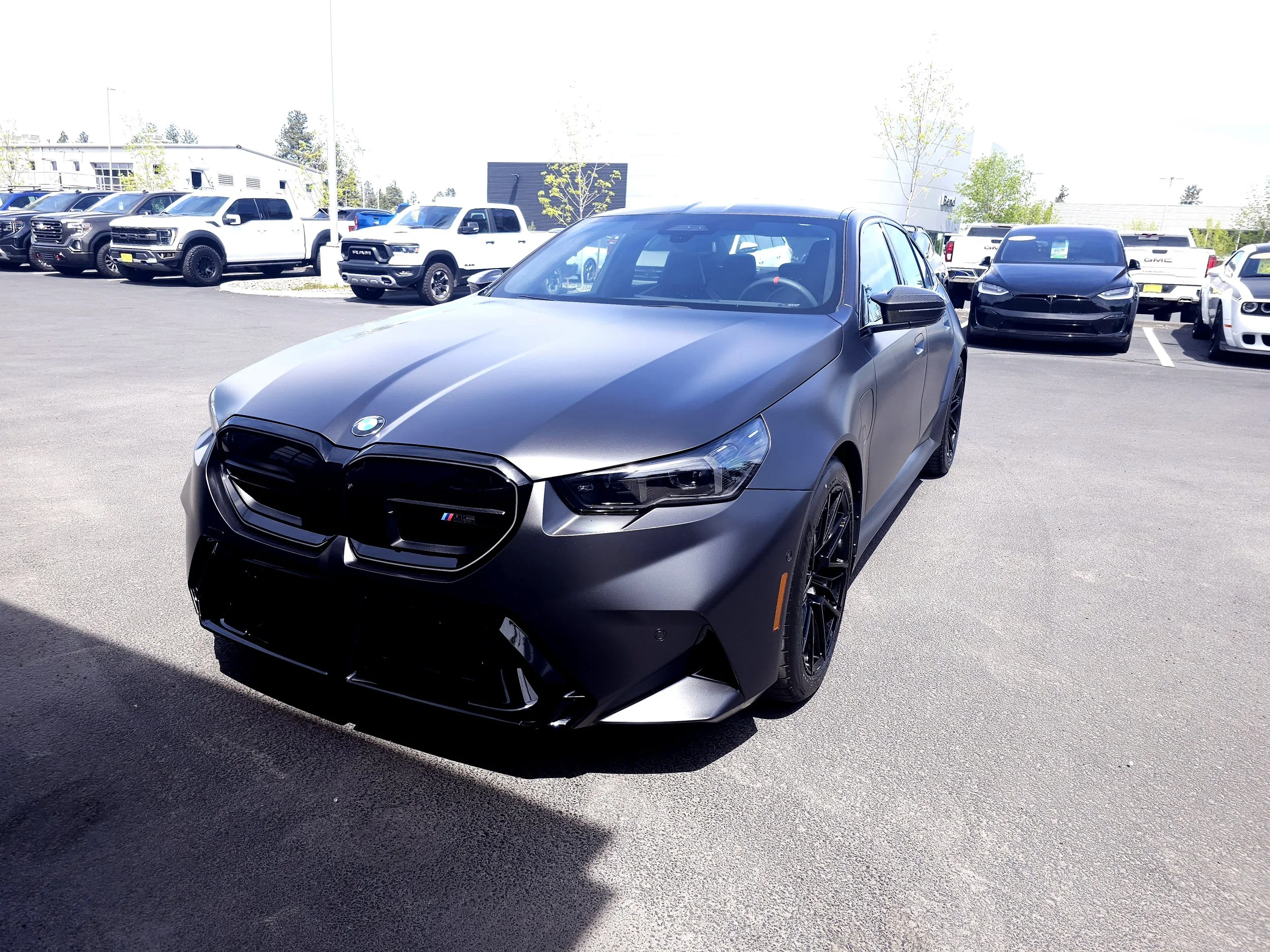 A row of parked vehicles, including a dark gray BMW M4 in the foreground, and other trucks and SUVs in the background, at a car dealership lot on a sunny day.