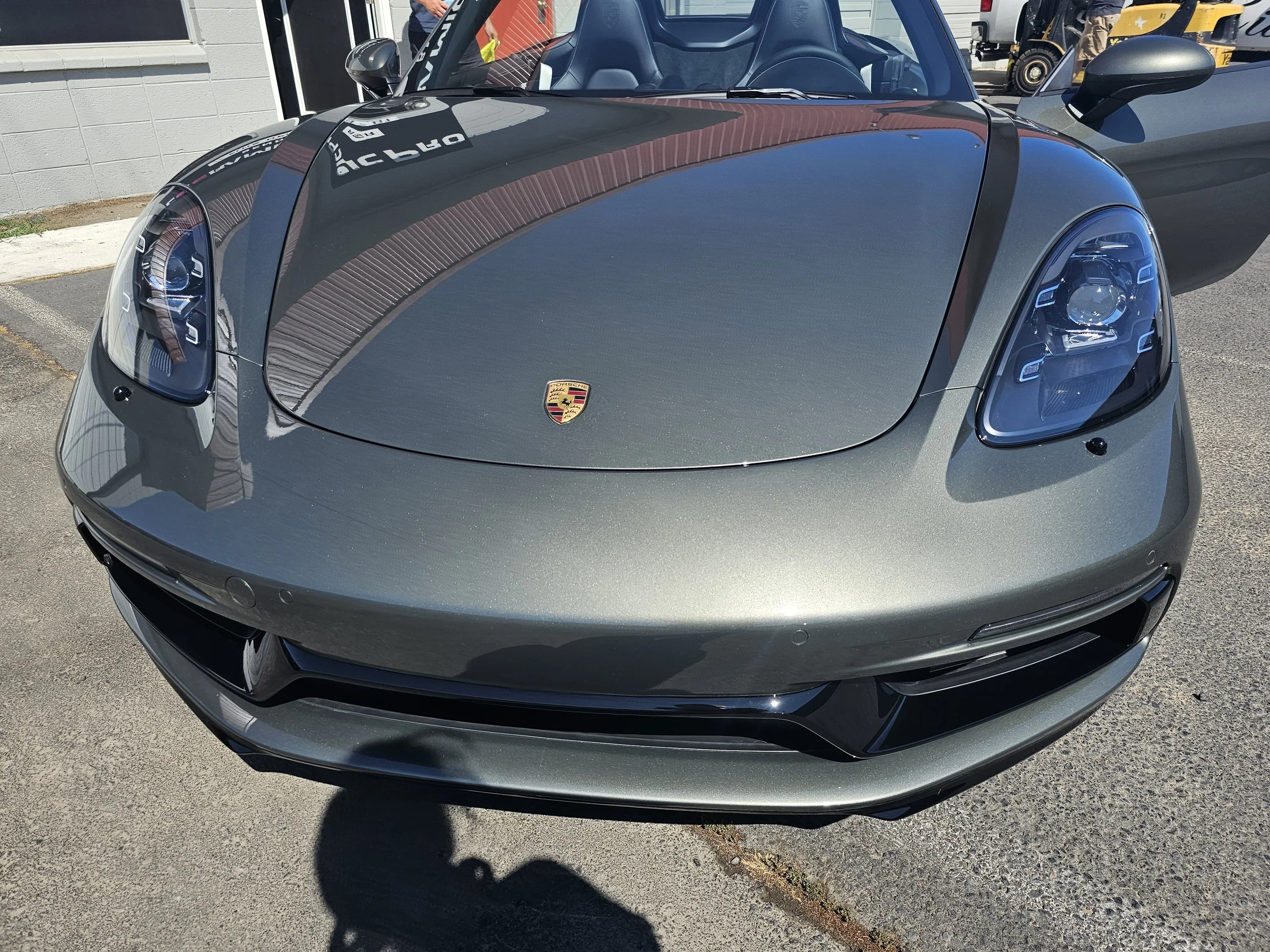 A sleek, gray Porsche sports car parked outdoors, with a partial view of its front end, including the hood, headlights, and windshield. The Porsche logo is visible on the front, and the car's reflections can be seen on its surface.