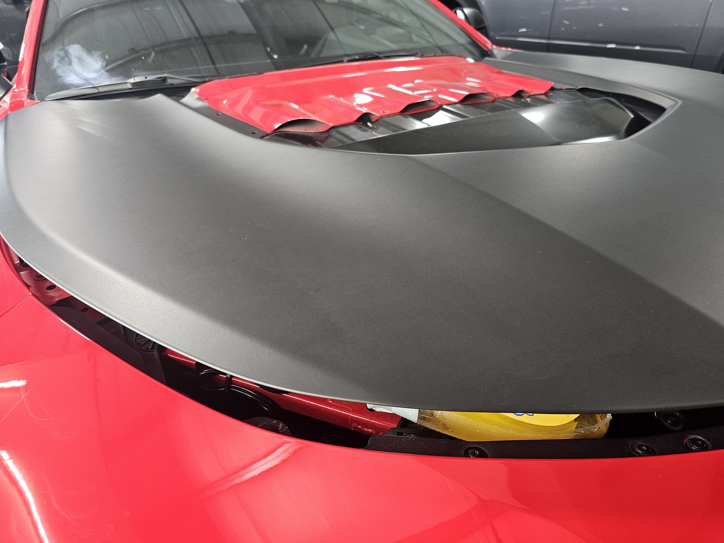 Close-up of the front of a red car with a black hood and a windshield, parked in a showroom or garage, with another dark-colored vehicle nearby.