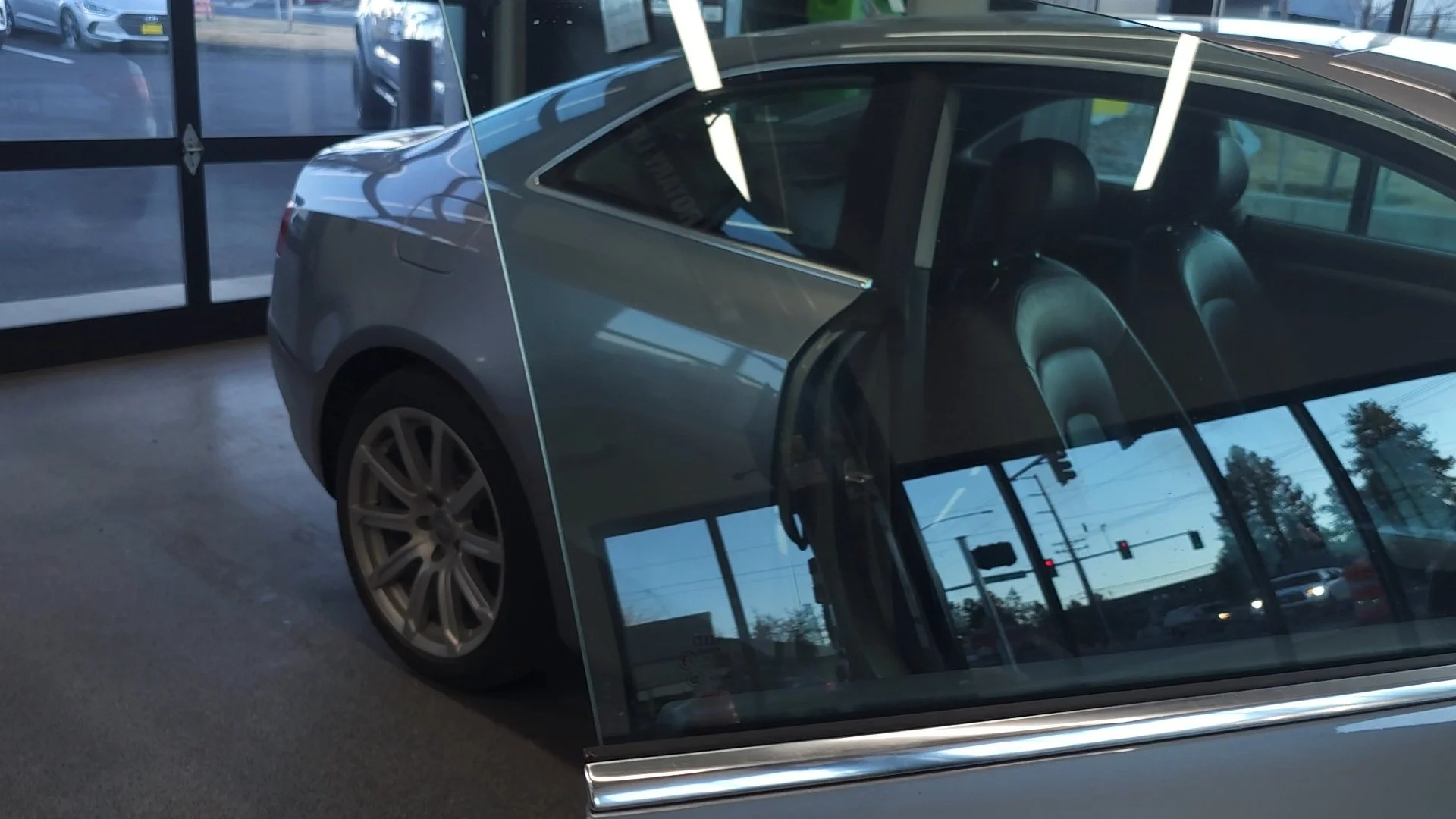 A gray car inside a showroom with a glass door. The car's reflection and interior seats are visible, along with the street outside and power lines.