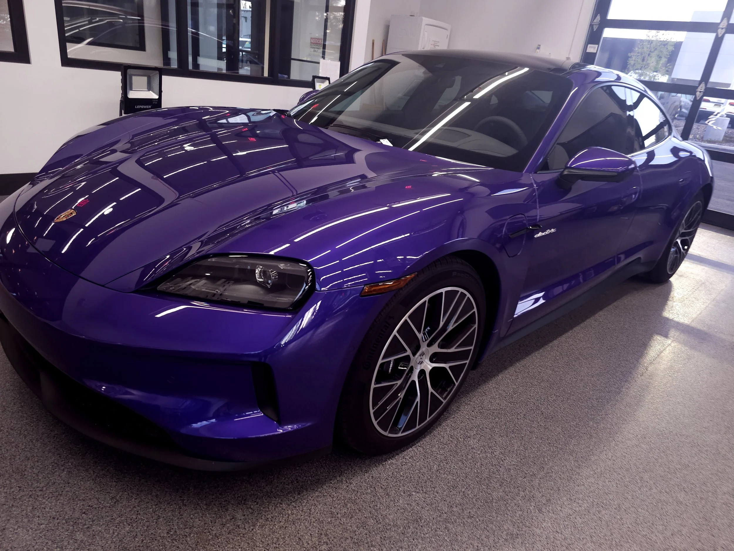 A purple sports car with a convertible top inside a car dealership or showroom, reflected by bright ceiling lights and large windows.