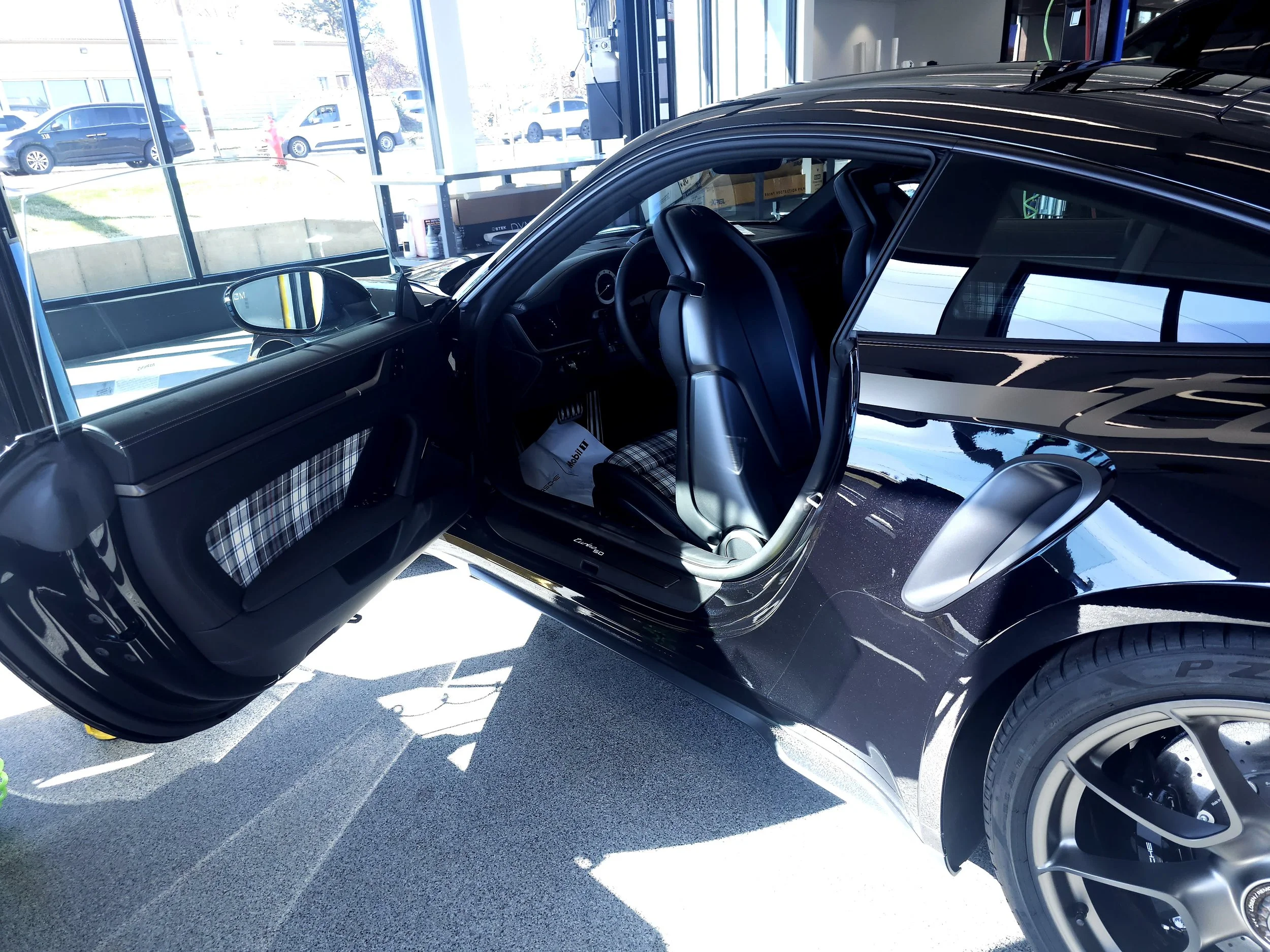 Black car with open door inside a dealership, showing plaid fabric seat covers and interior; outside visible through windows with parked cars and a hose in the background.