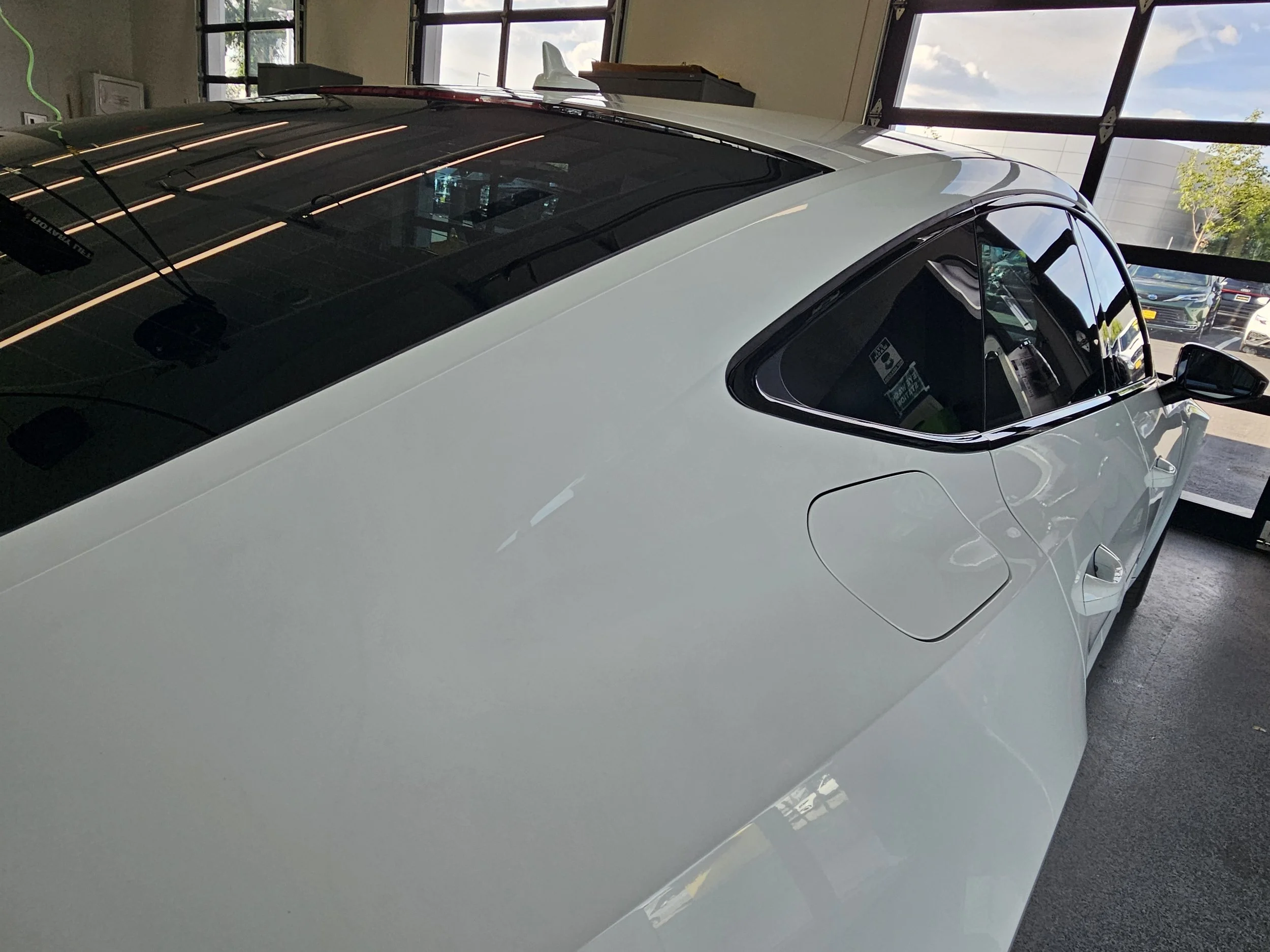 A white Tesla sports car with a black roof inside a showroom or dealership, with large windows showing the parking lot and trees outside.