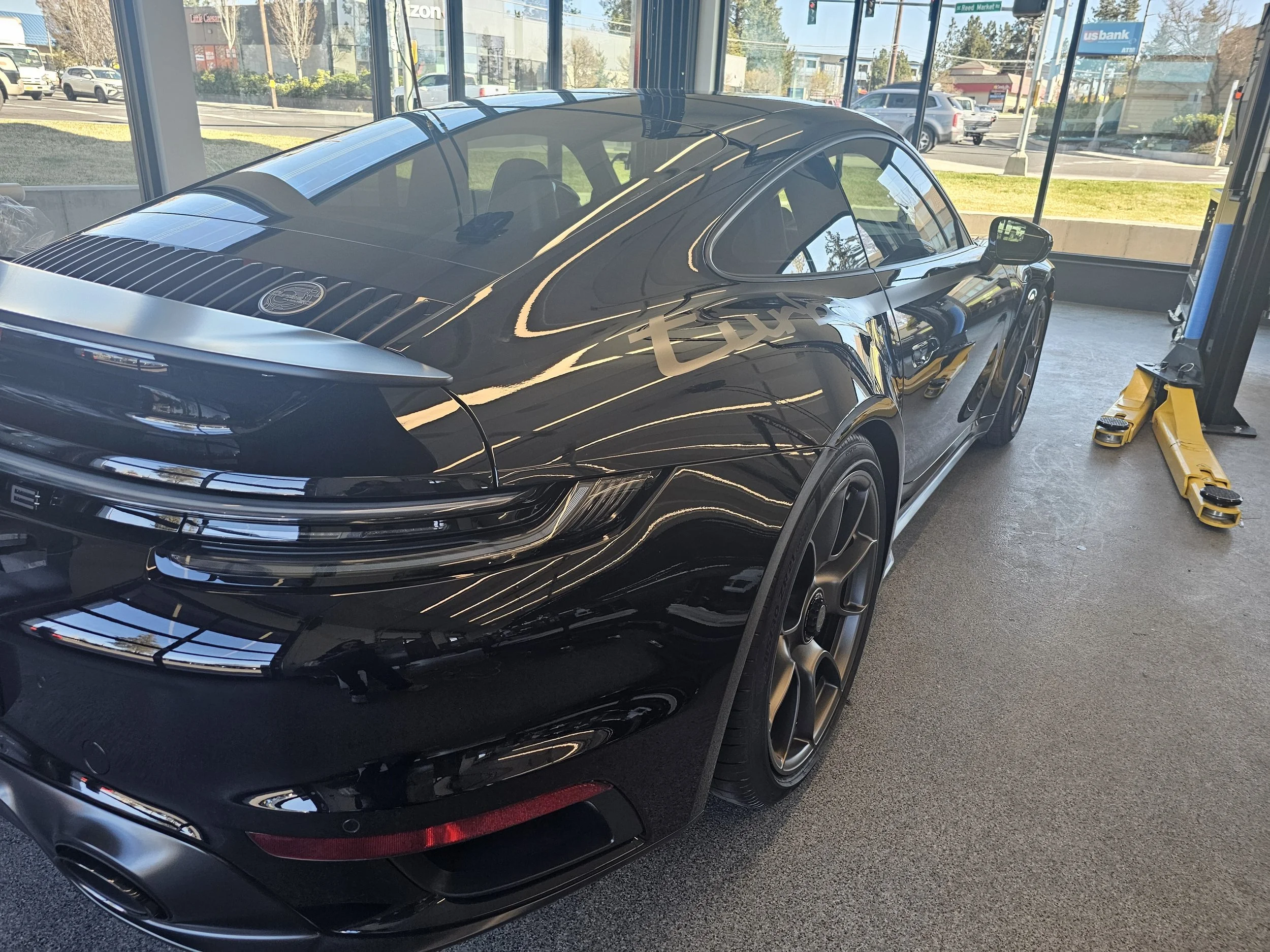 A black Tesla Model X parked inside a car dealership or service center, with a yellow hydraulic lift on the right side and large glass windows showing a street with parked cars and buildings outside.